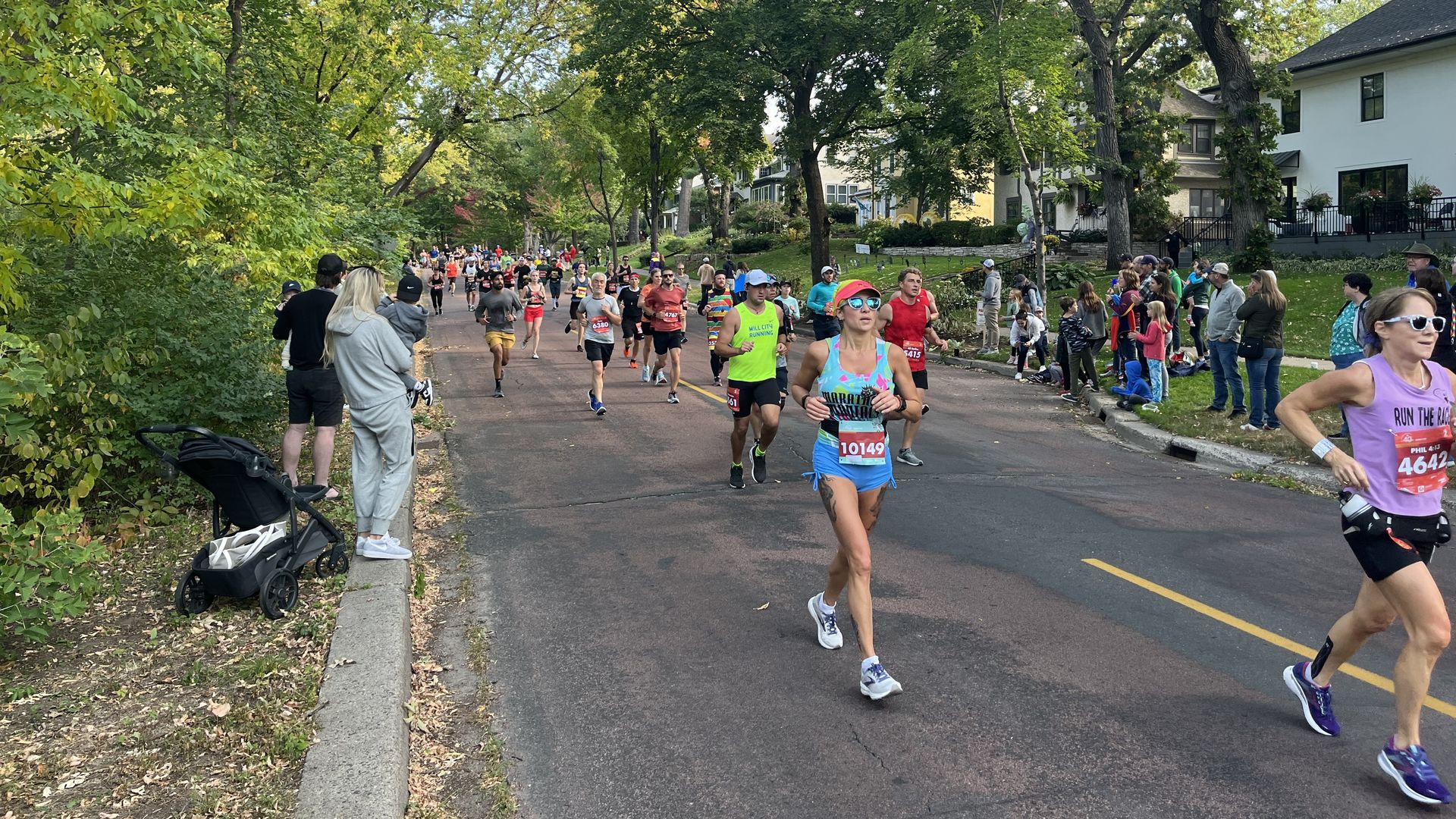 Runners in a city marathon on a tree-lined street; spectators line the sidewalk by white houses. A stroller sits on the left as runners pass by in bright athletic gear.