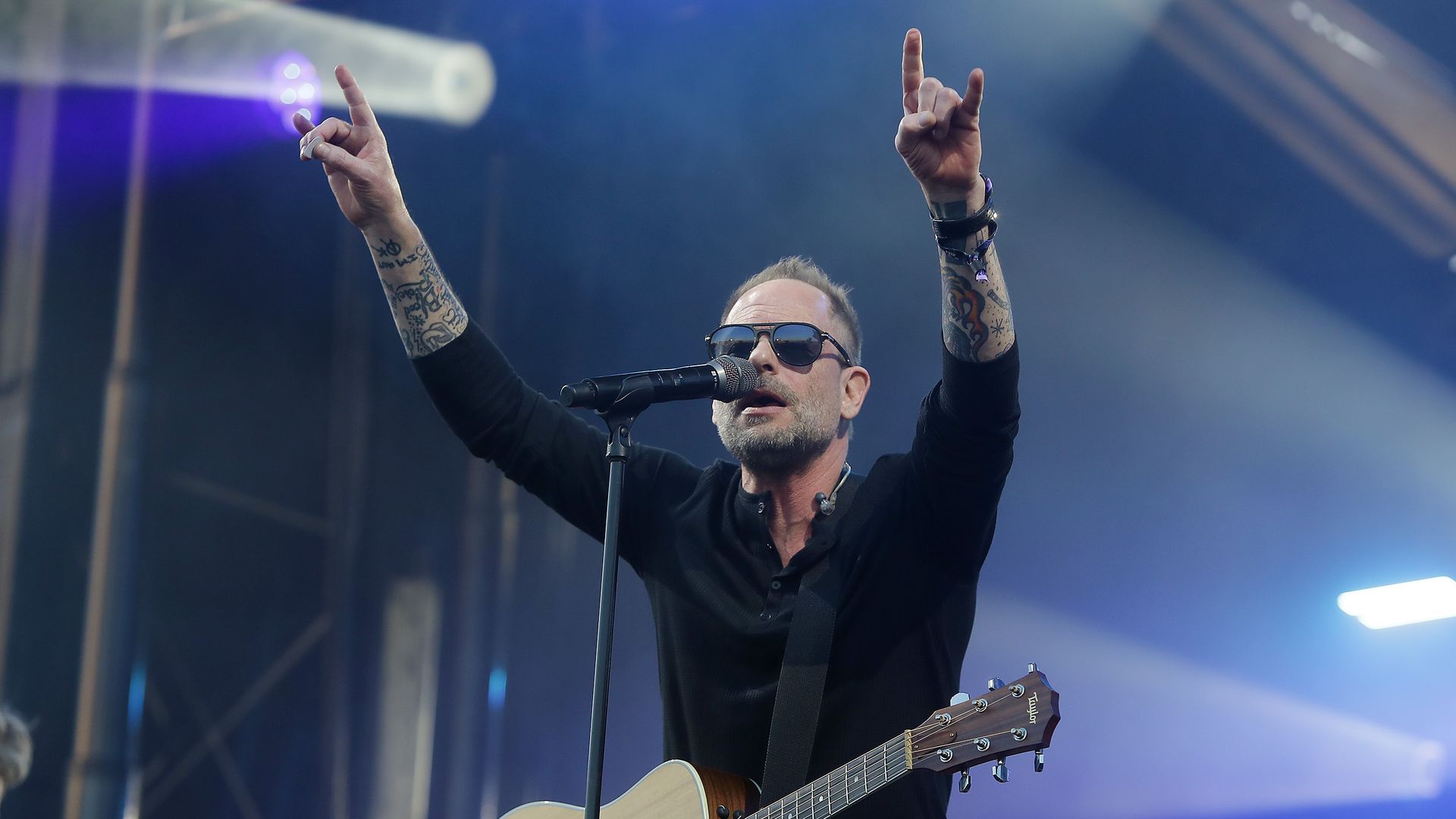 Male musician with tattoos and sunglasses singing into a microphone, playing an acoustic guitar, raising both hands in a rock gesture on a blue-lit stage.