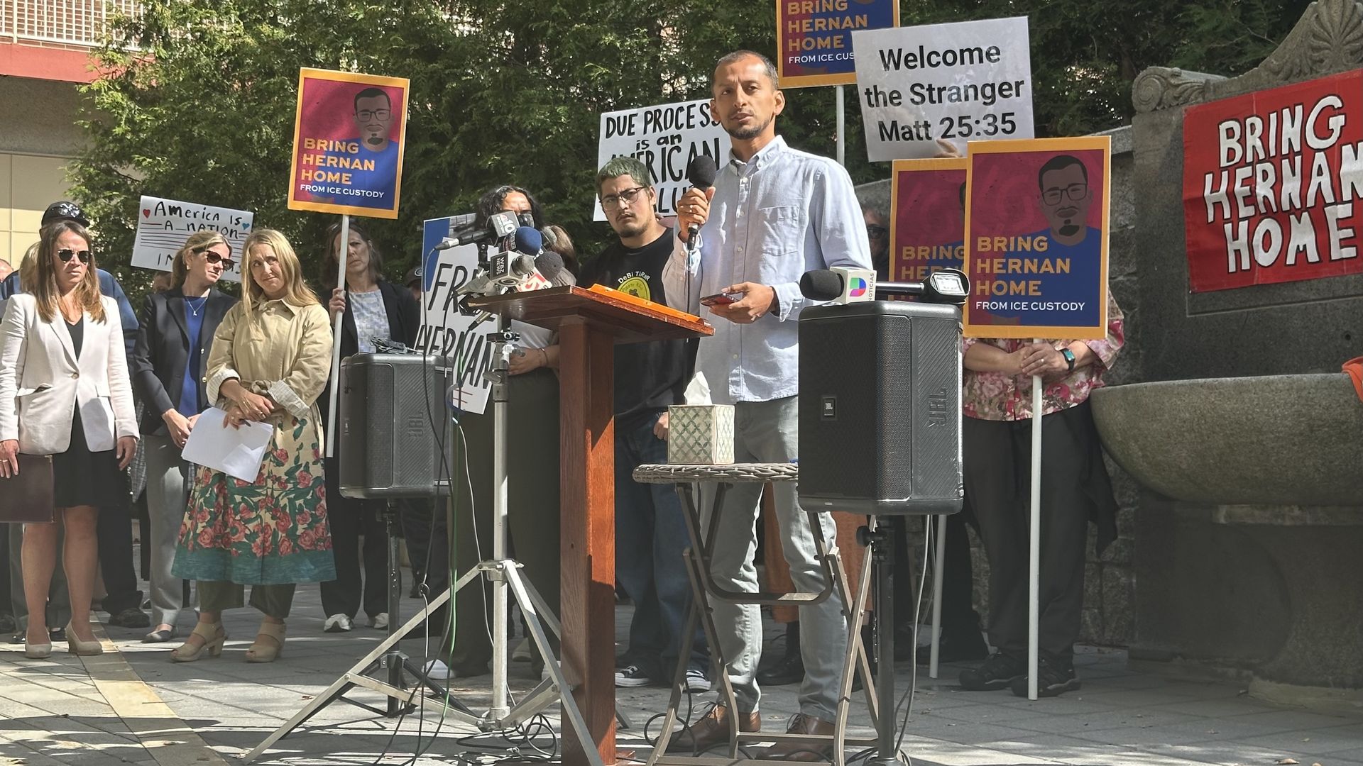 Frank Pinto, a friend of detained Edgar Hernan Elias Escobar and his wife Leslie Perlera Gonzalez, speaks at a lectern holding up a microphone surrounded by people calling for Hernan's release from ICE custody. 