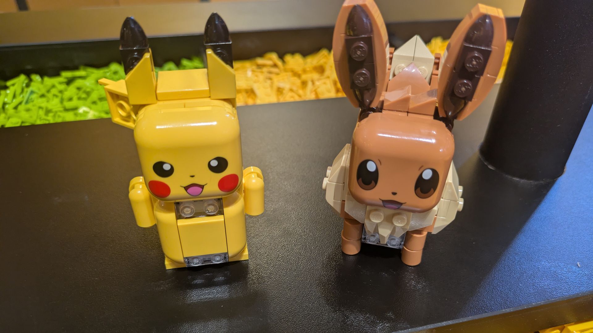 Two LEGO animal figures on a black surface: a yellow Pikachu-like figure with black ears on the left, and a brown deer-like figure with big ears on the right, set against yellow and green bricks.