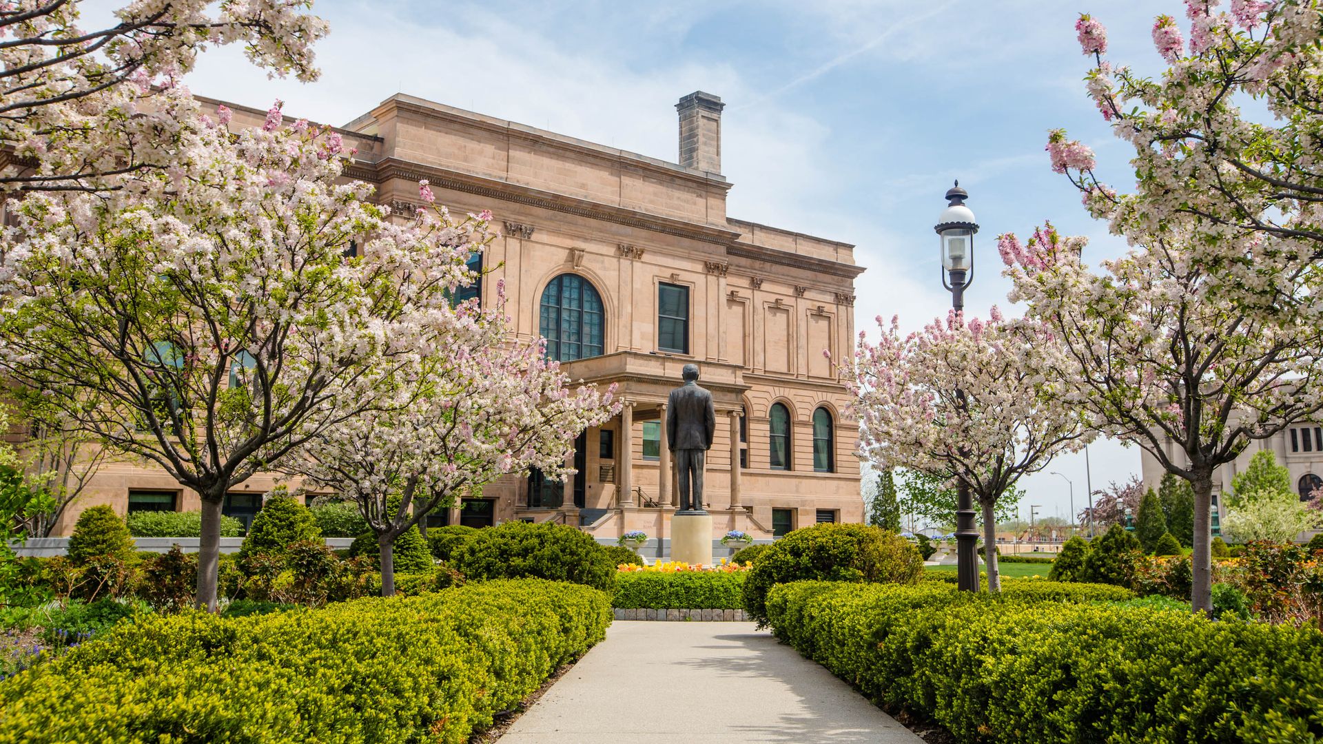 Campus building in warm stone framed by blooming cherry trees with pink and white blossoms. A statue stands along a paved path, bordered by hedges and a lamppost under a blue sky.