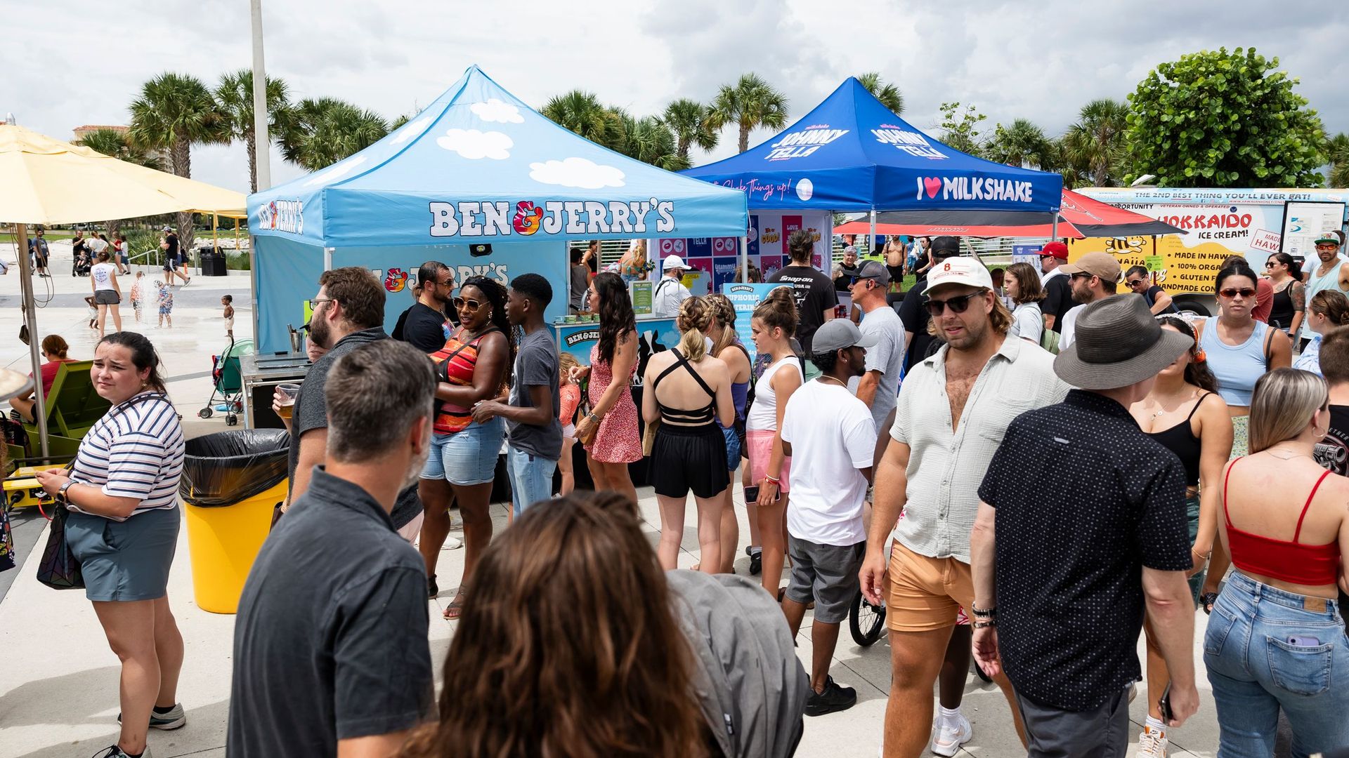 A couple dozen people walking by a light blue tent decorated with clouds that says "Ben & Jerry's" and a royal blue tent that says "I <3 milkshake."  