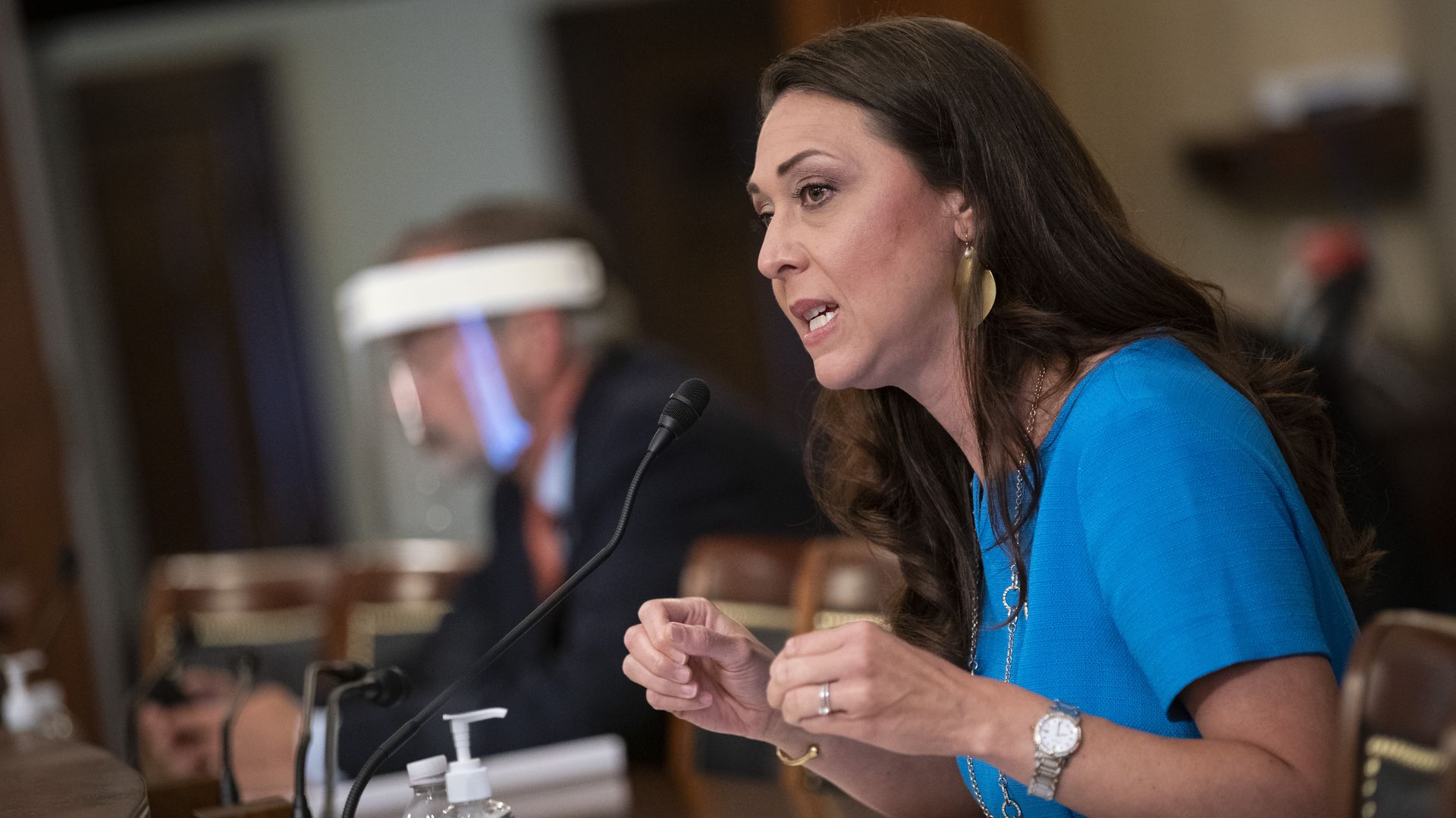 A woman with brown hair dressed in blue gestures with her hands while speaking. 