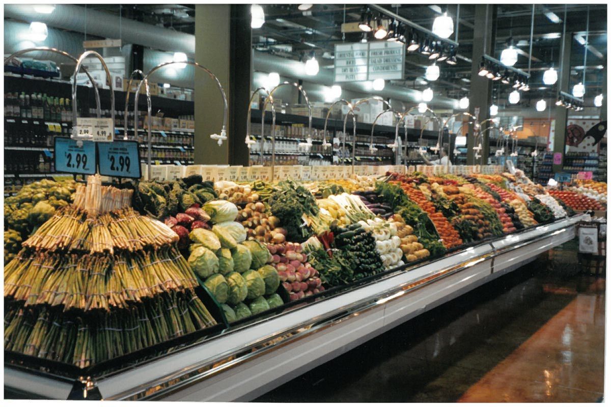 Supermarket produce section with neatly stacked asparagus, cabbage, onions, leafy greens, carrots, and a variety of vegetables under bright overhead lights and price signs.