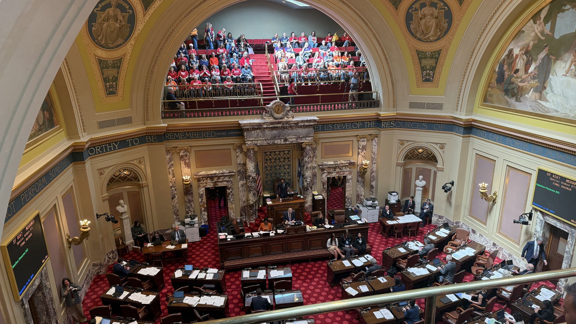 Interior of a grand legislative chamber with a red carpet and desks; a balcony gallery filled with people in red shirts, ornate arches, gold trim, and murals.