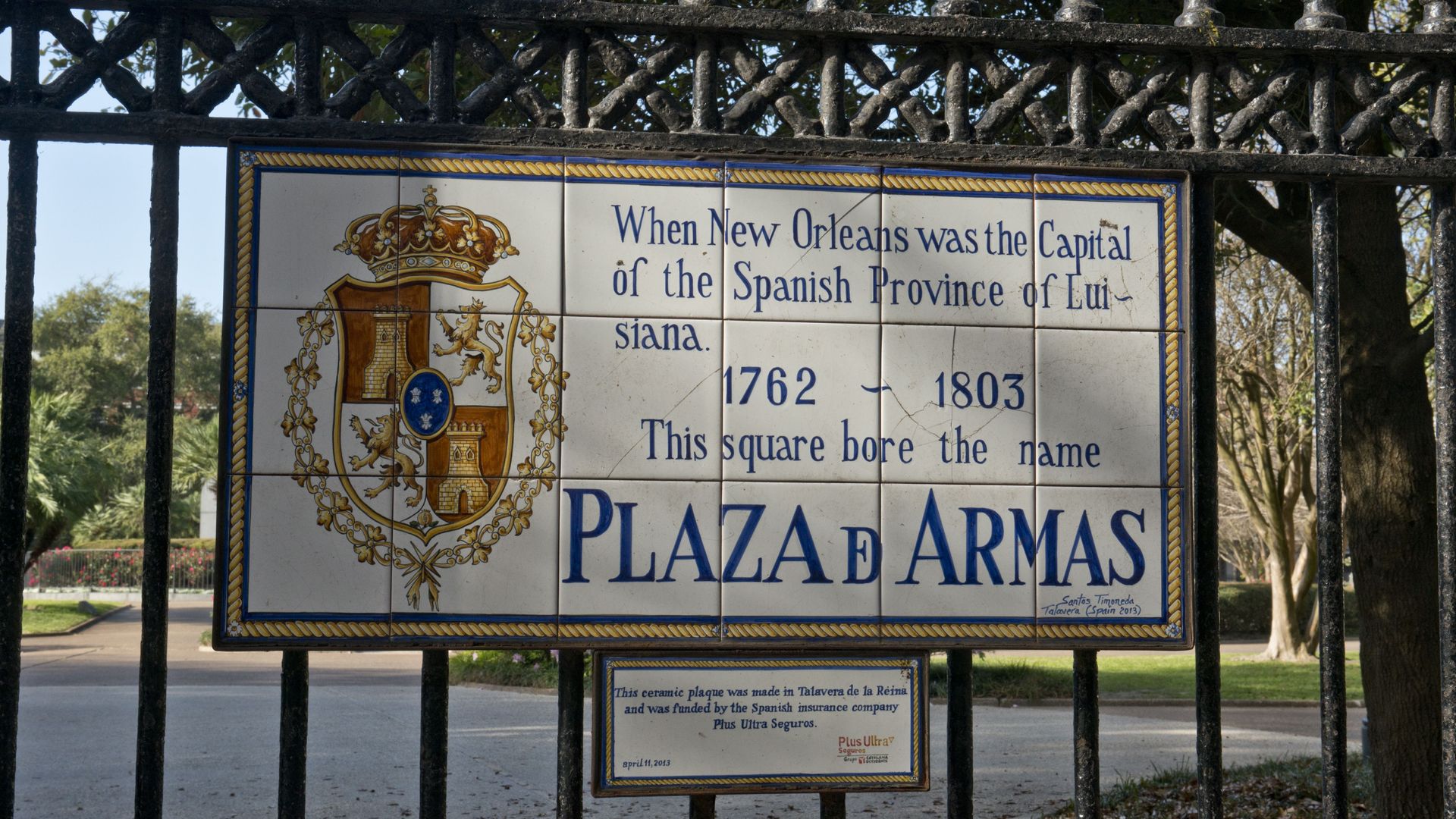 A tile sign hangs on a fence and says "When New Orleans was the capital of the Spanish province of Luisiana, this square bore the name Plaza D'Armas."