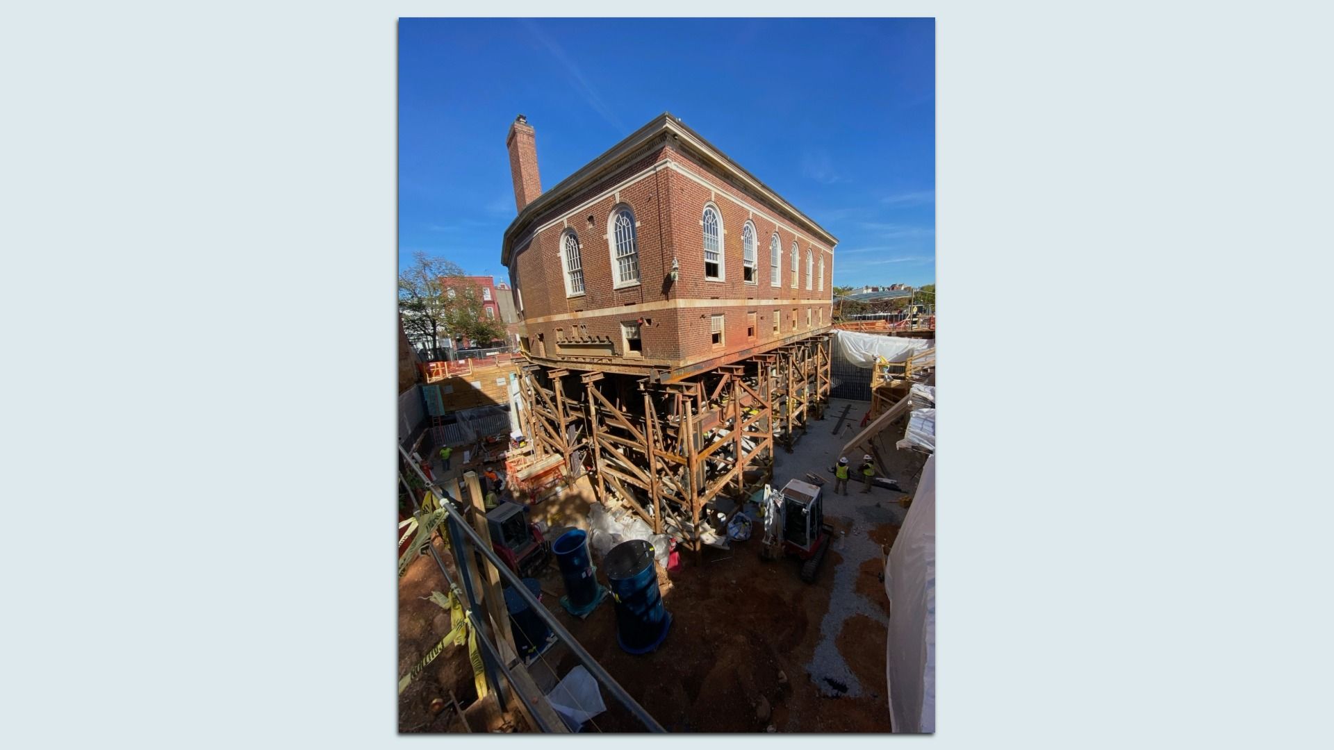 Historic brick building with tall arched windows perched on extensive wooden scaffolding at a construction site. A tall brick chimney rises to the left; clear blue sky above, workers below.