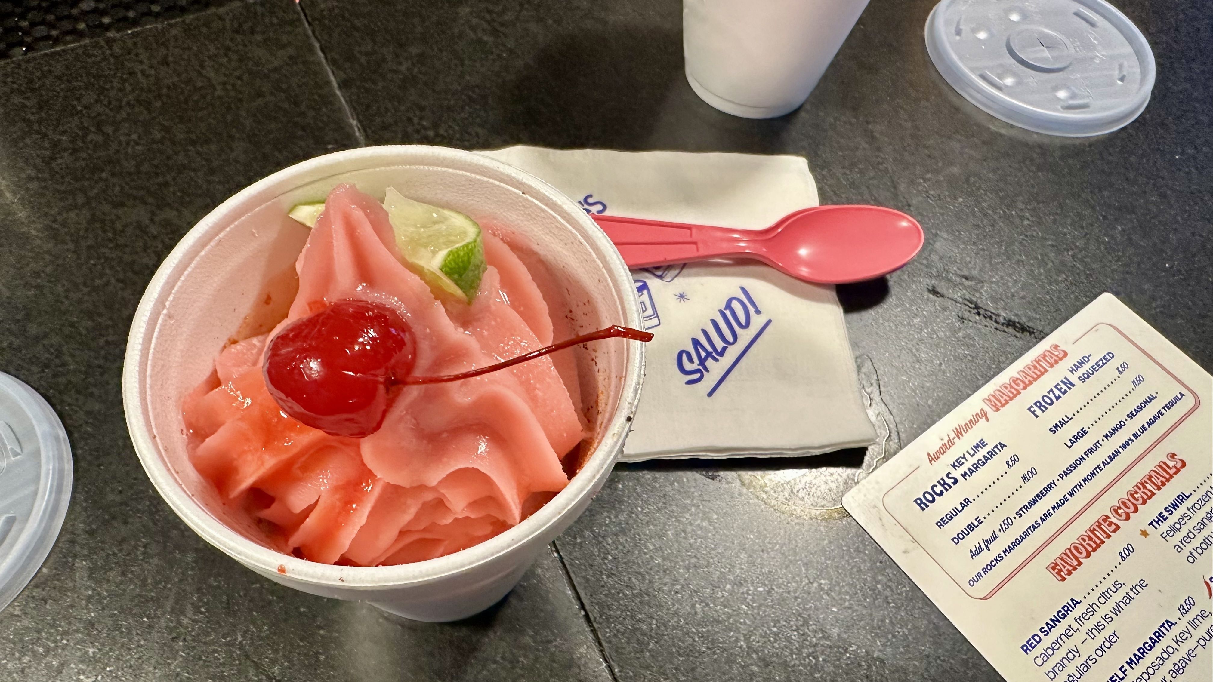 Pink frozen dessert in a white foam cup topped with a red cherry and lime wedge; a bright pink spoon rests on a napkin beside a white takeaway cup on a dark counter, with a menu nearby.