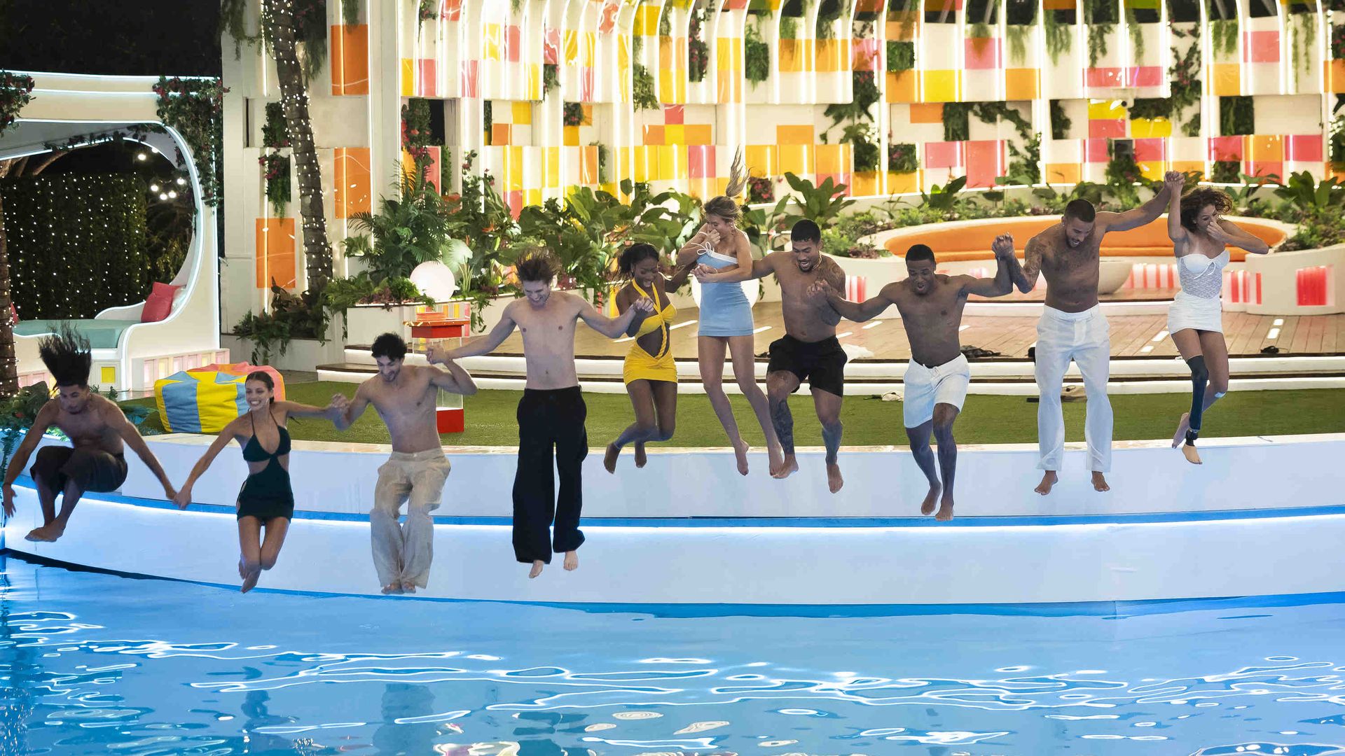 Diverse group jumps into a bright resort pool at night, holding hands in a line. Colorful tiled wall, string lights, tropical plants, and a curved white seating alcove on the left.