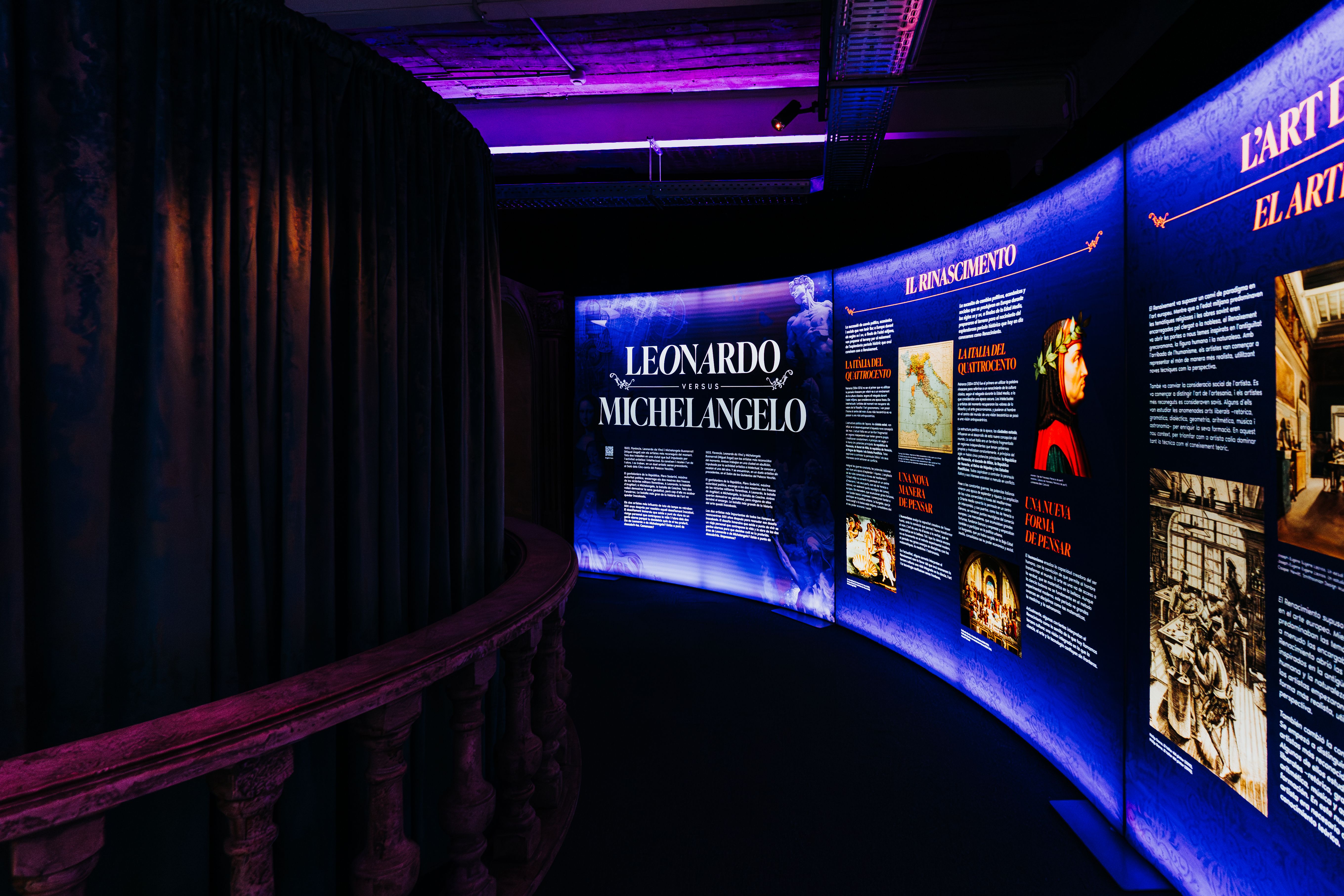 Dimly lit museum corridor with dark curtains on the left and a curved railing. Blue-lit exhibition boards show the title "LEONARDO VERSUS MICHELANGELO" and "IL RINASCIMENTO" with images and text.