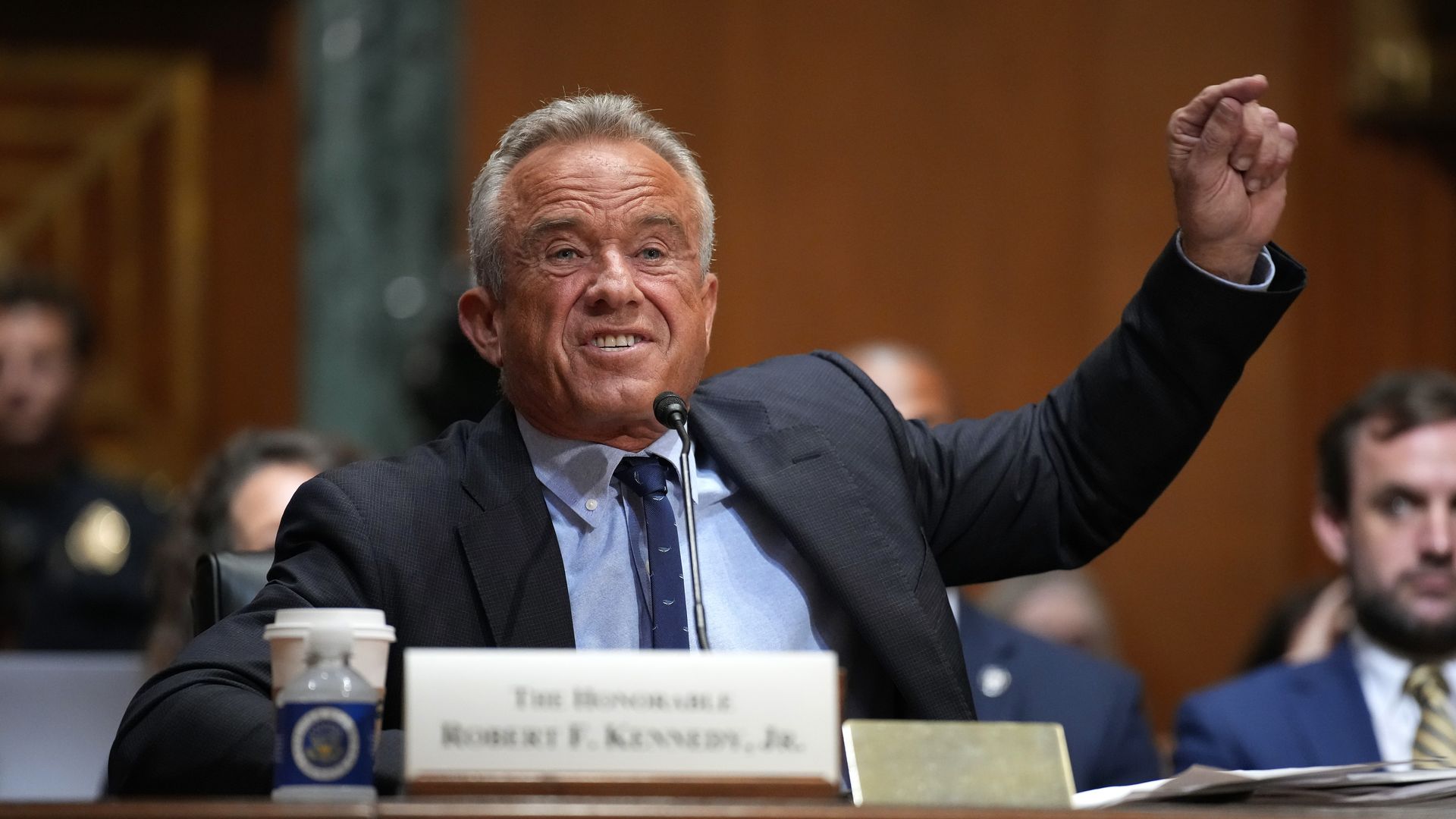 RFK Jr., wearing a dark suit and a blue shirt, raises his arm and points while speaking at a hearing before a Senate panel (lawmakers are not pictured).