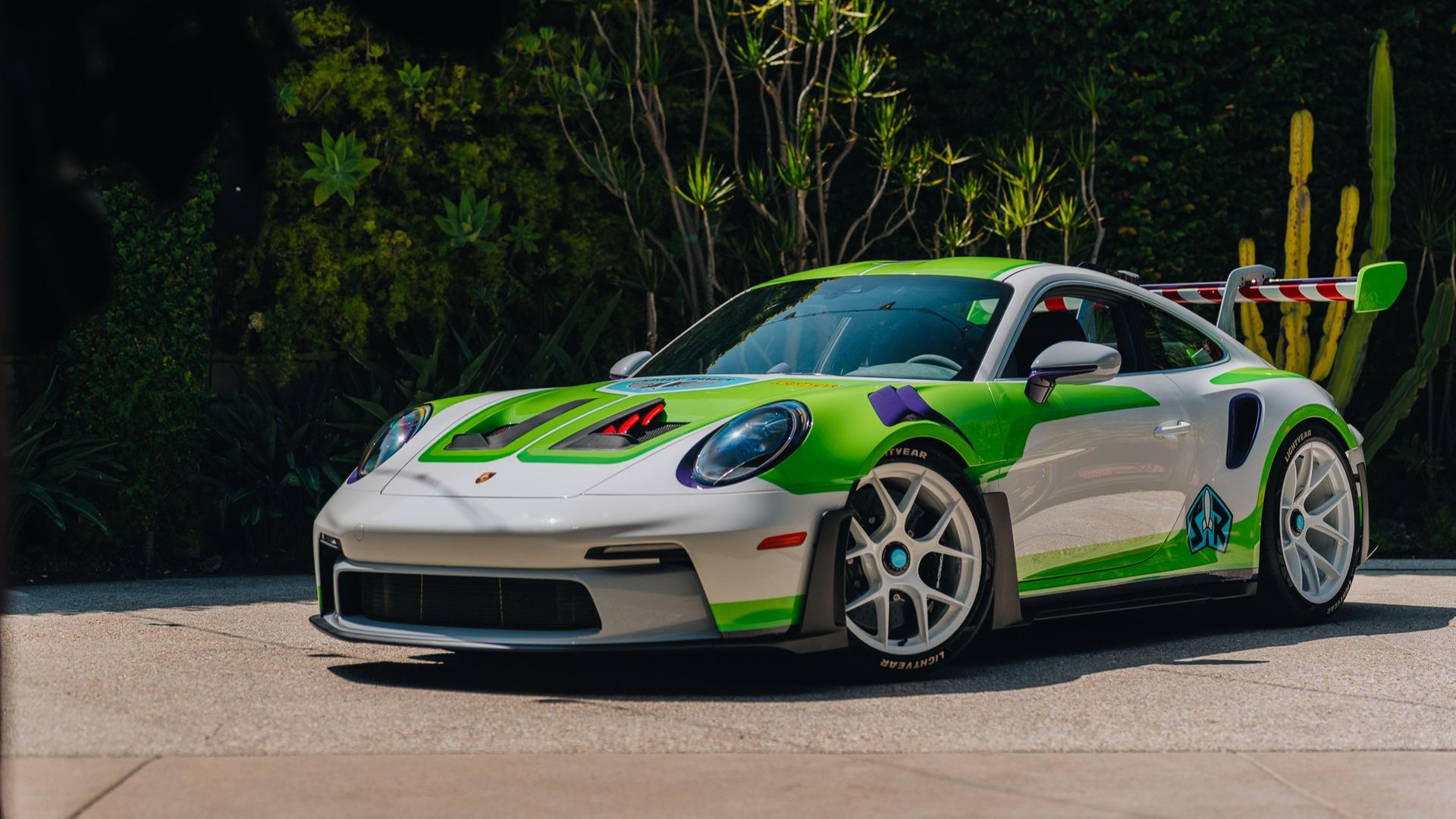 A white Porsche race car with lime-green accents and a large rear wing sits parked on a sunny pavement, with lush tropical plants and tall cacti in the background. The car resembles Buzz Lightyear from Toy Story