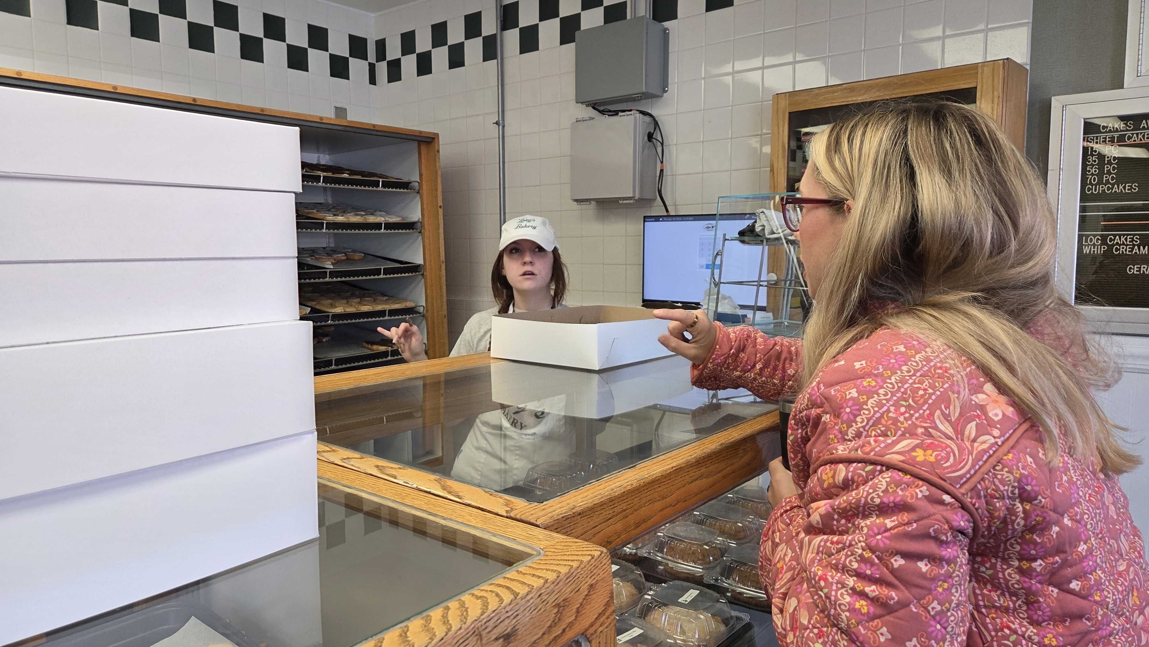 Bakery counter scene: customer in a pink floral jacket stands at a glass display as a staff member wearing a white cap hands over a box; pastries line shelves behind white tiled walls.