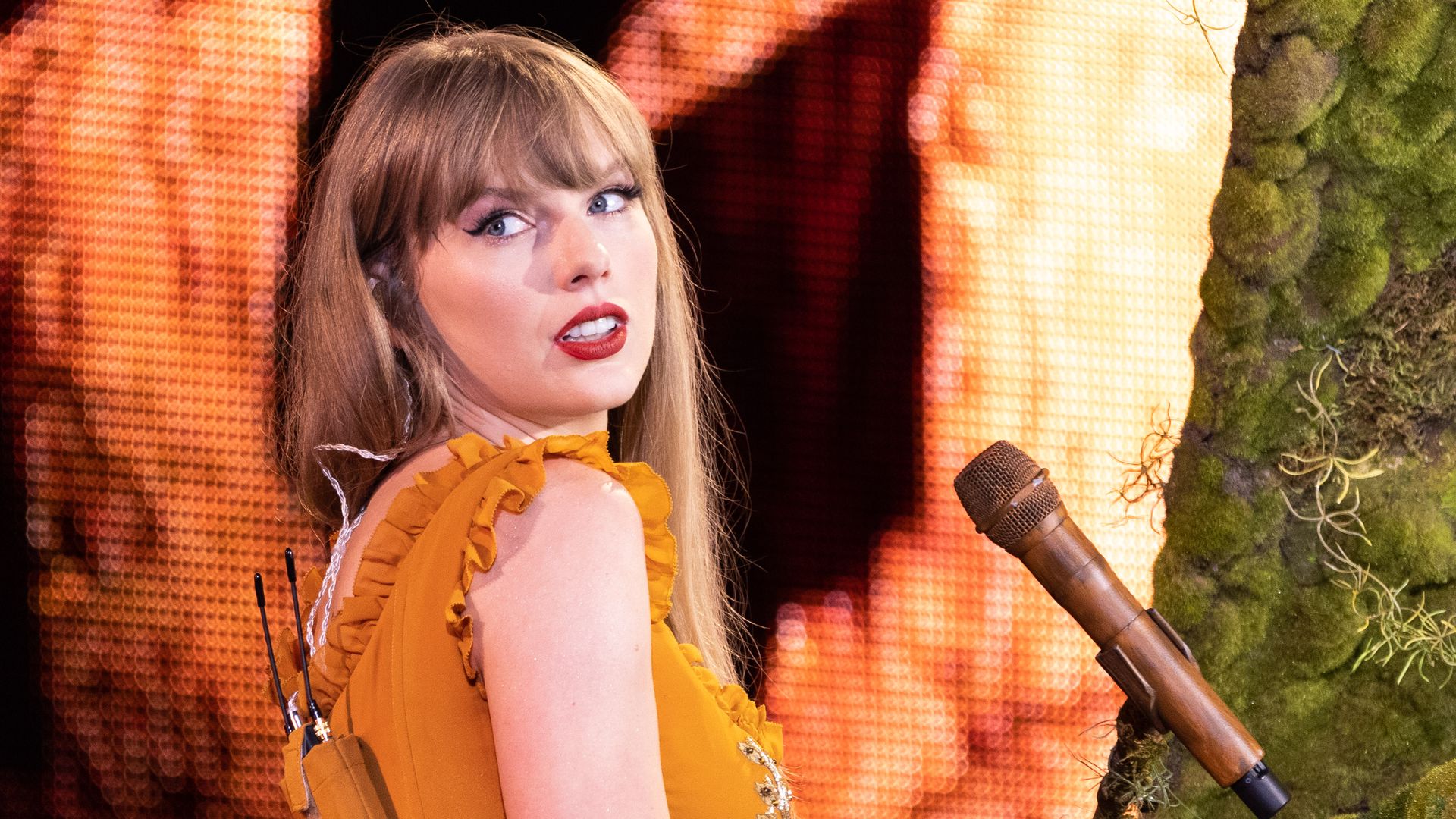 An individual wearing an orange dress while performing at a piano covered in moss looks over their shoulder