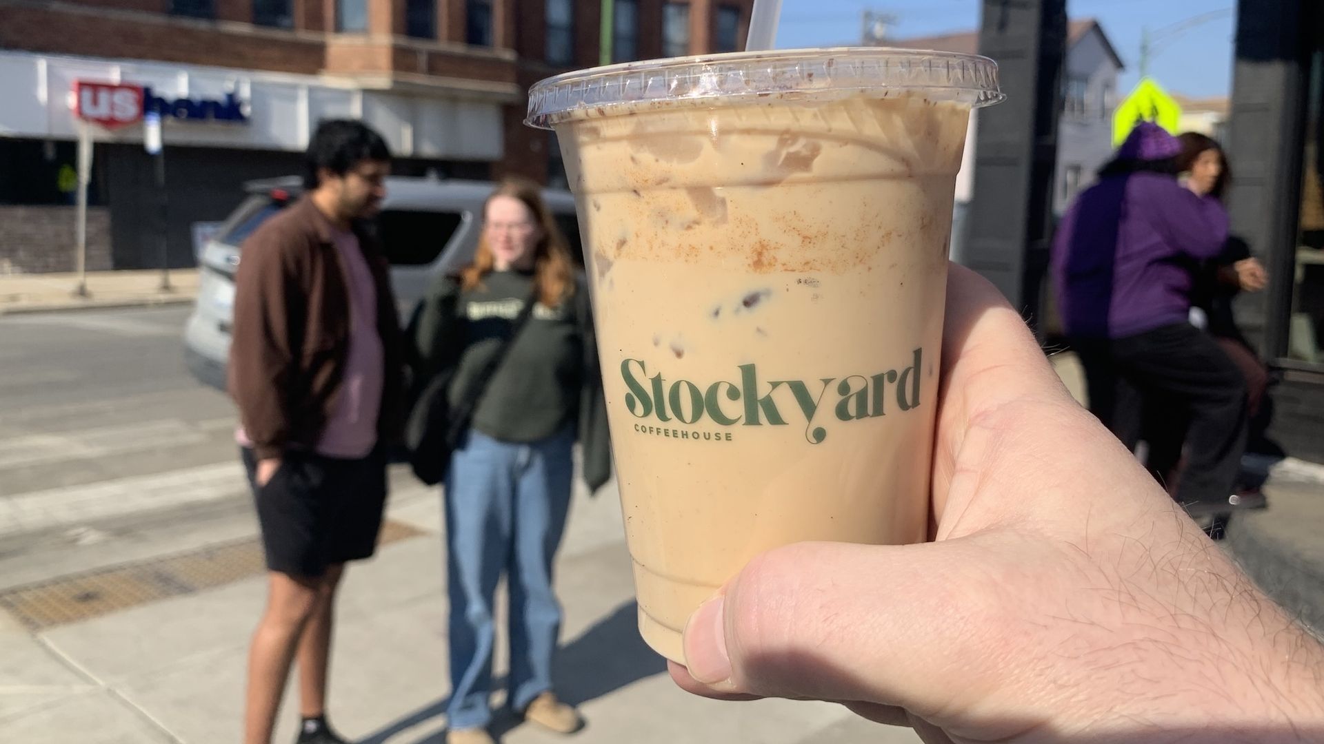 Hand holding an iced coffee cup from Stockyard Coffeehouse outdoors on a sunny day, with blurred people and buildings in the background.