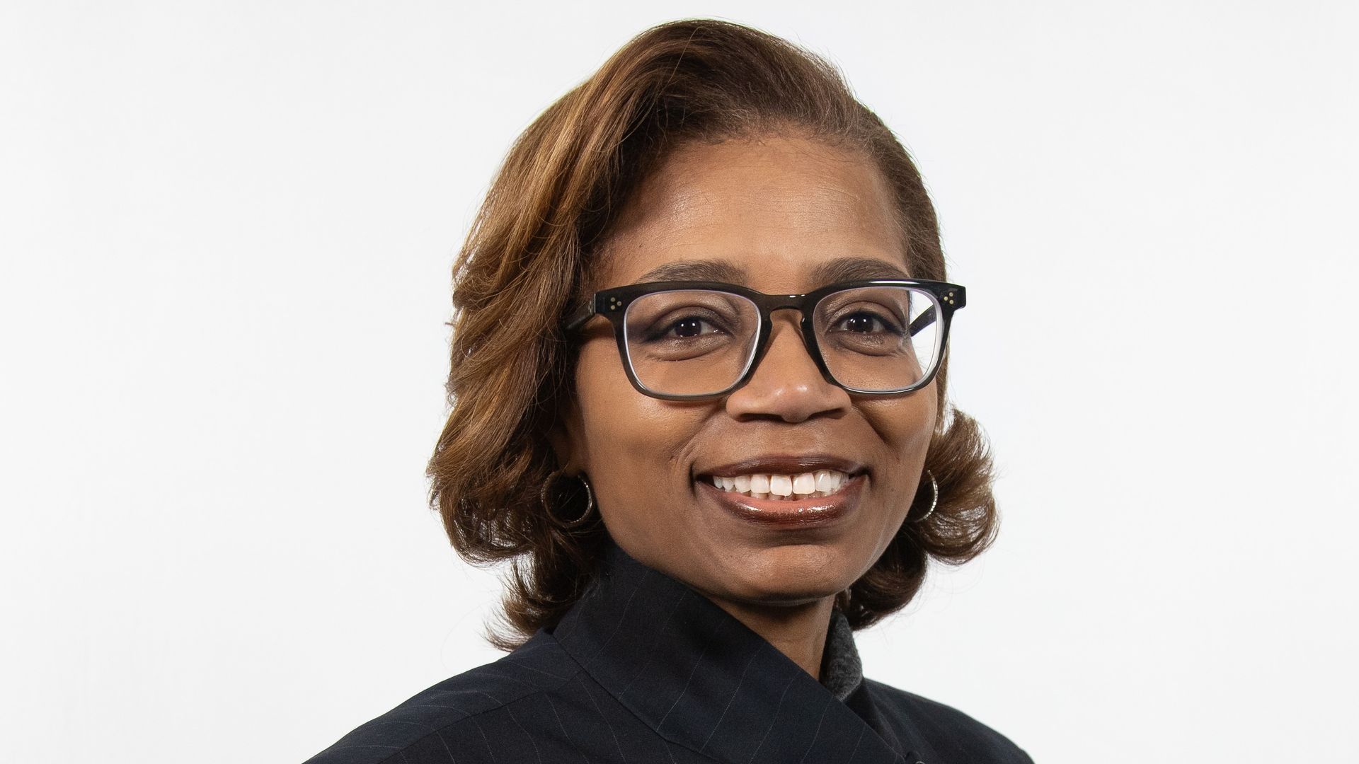 Portrait of a smiling Black woman wearing black-framed glasses and a dark pinstriped blazer, with short brown hair, looking at the camera against a white background.