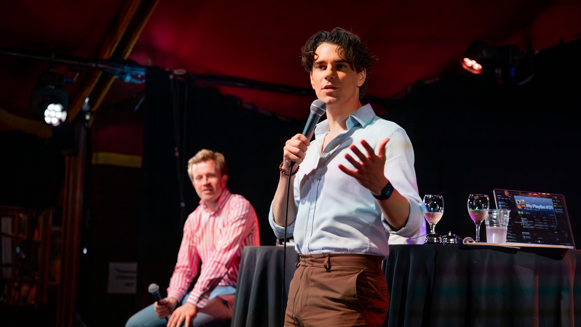 Two men on stage at a talk or podcast event; one in a white shirt and brown pants is speaking into a microphone, the other in a red striped shirt and jeans is seated holding a microphone.