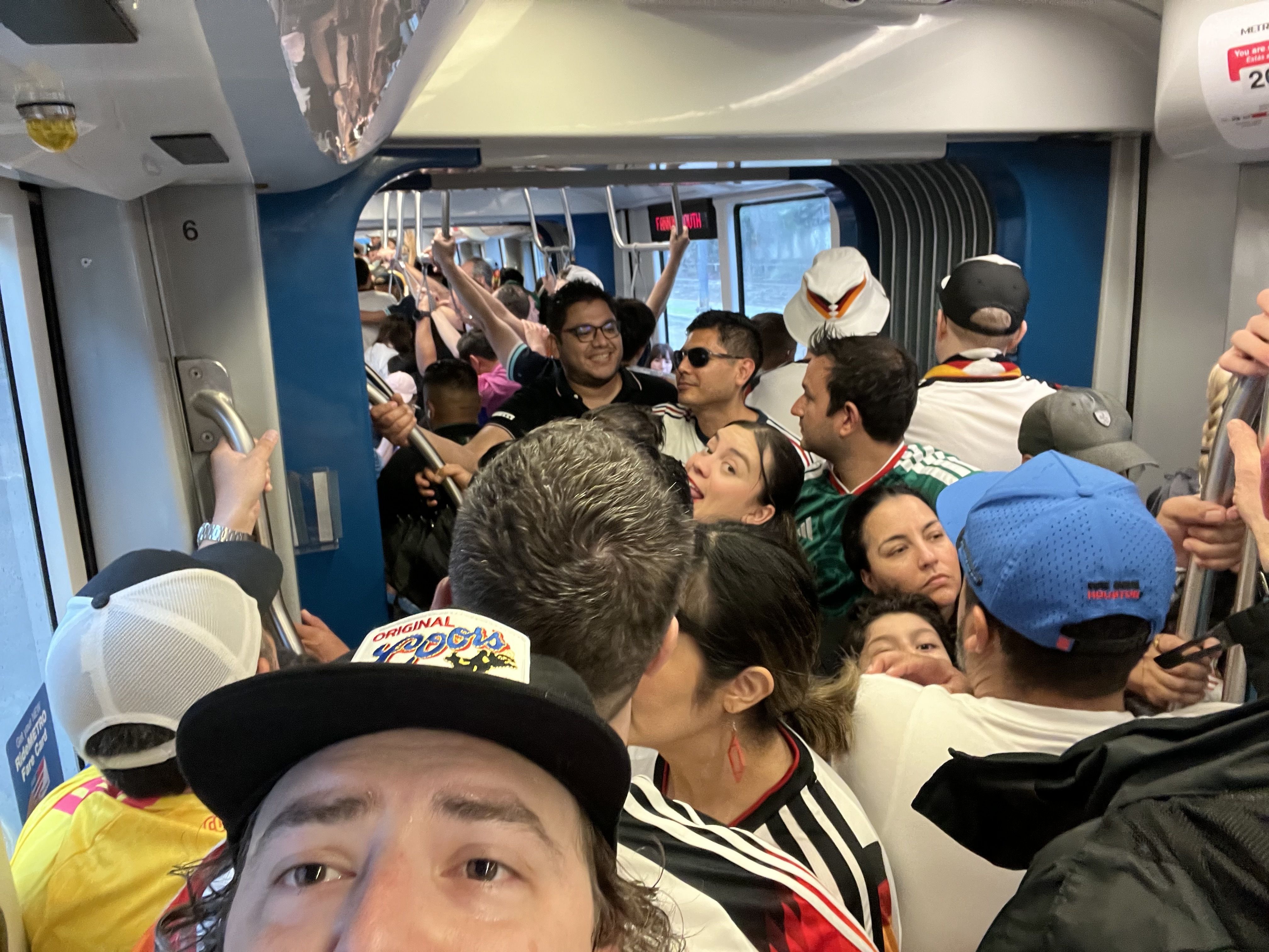 Inside a packed subway car, riders stand shoulder to shoulder, gripping overhead straps and poles. They wear colorful shirts, hats, and sunglasses as blue walls frame the crowded scene.