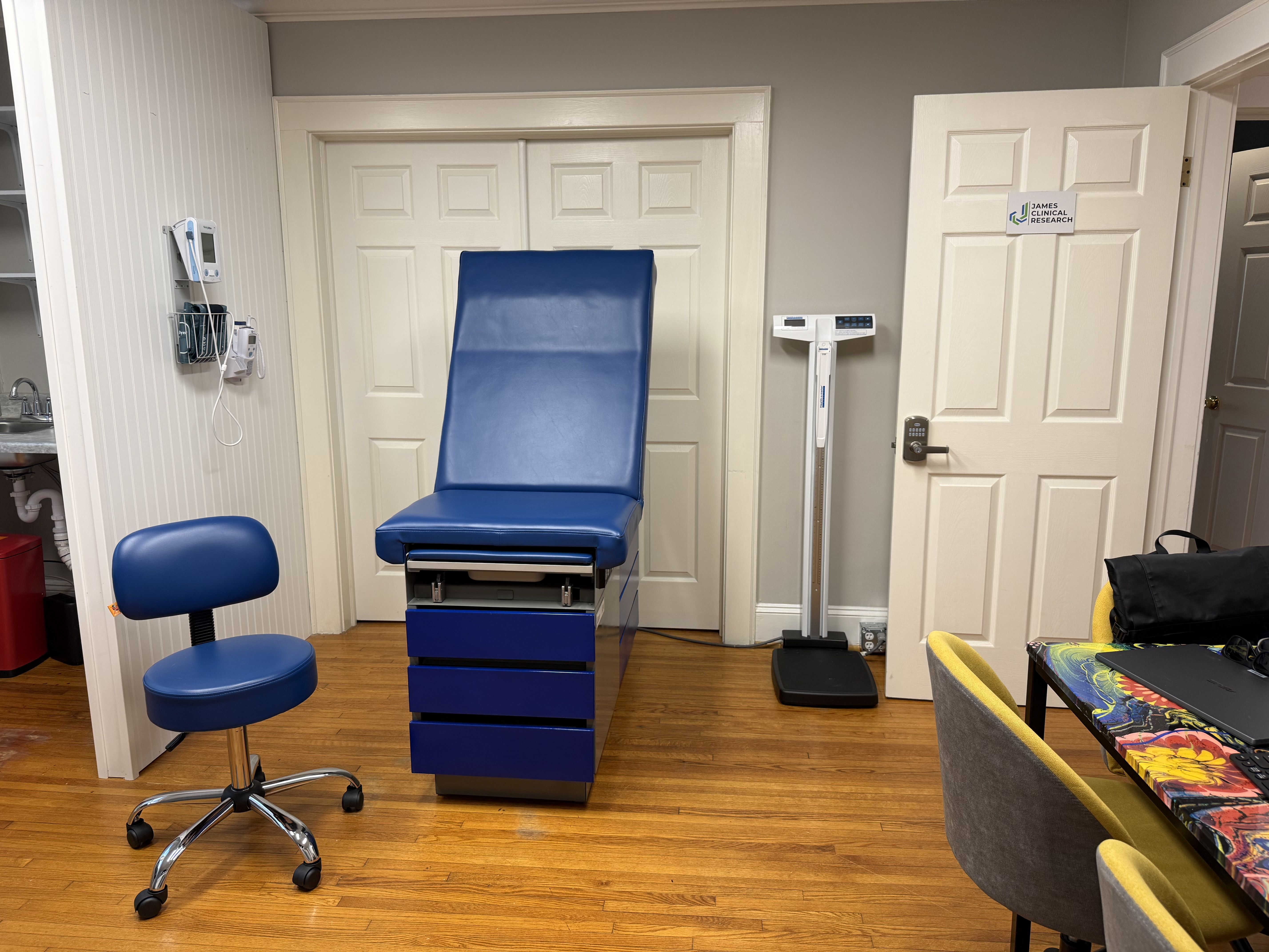 Medical exam room with a tall blue chair, matching stool, wall-mounted blood pressure monitor, scale, white paneled doors, gray walls, and a sign reading James Clinical Research on the door.