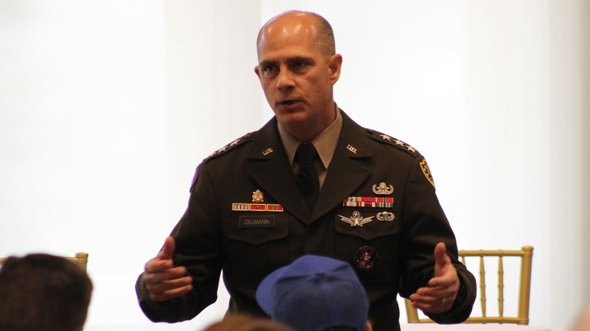 A bald man in a U.S. Army dress uniform speaks at a conference, showing ribbons and medals. His name tag reads Zellmann as he gestures to the audience.