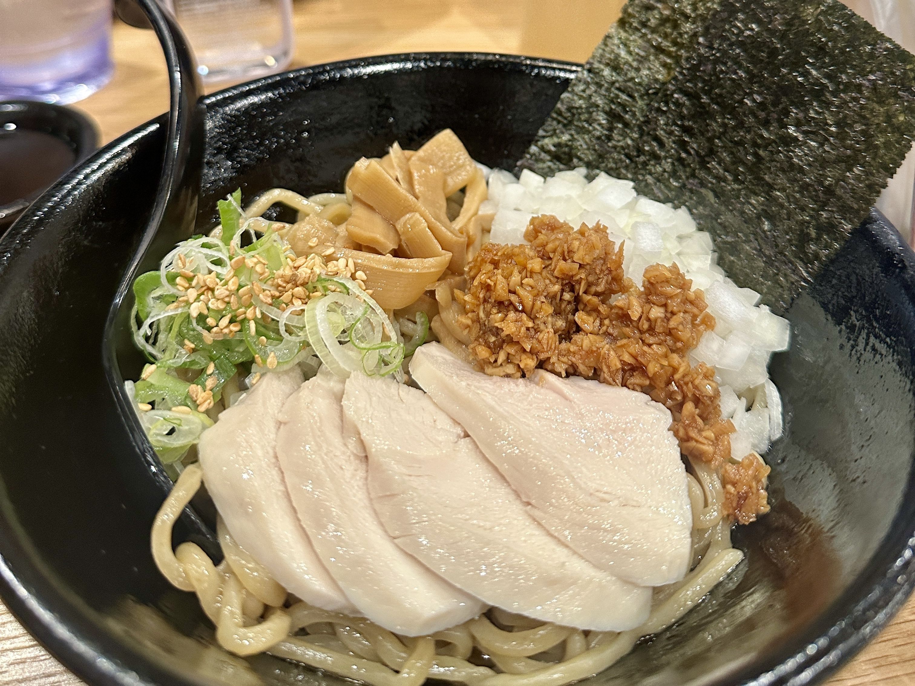 Close-up of a black ramen bowl with sliced chicken, bamboo shoots, diced onions, chopped green onions with sesame seeds, nori sheet, all on noodles.