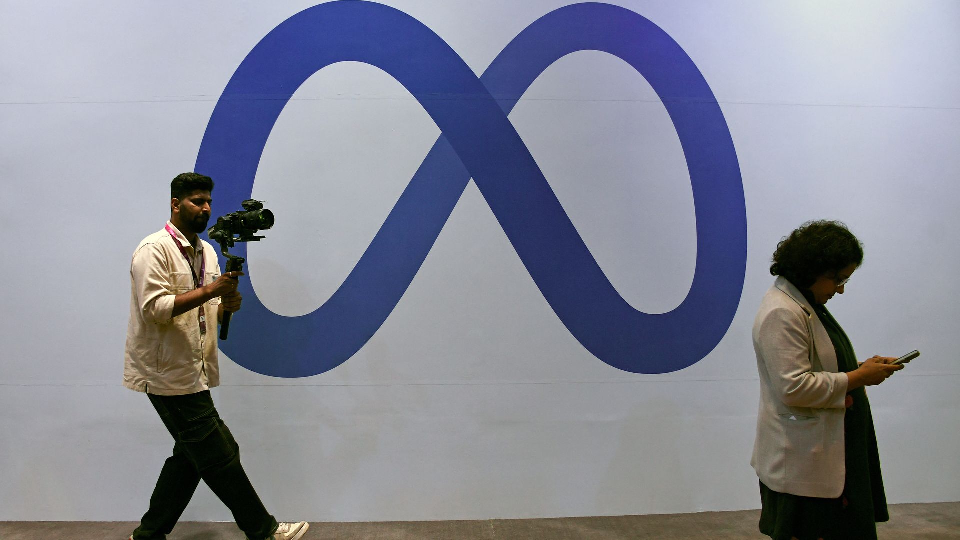 Man holding a camera walks in front of a large blue Meta logo on a white wall, while a woman in a beige jacket looks at her phone nearby.