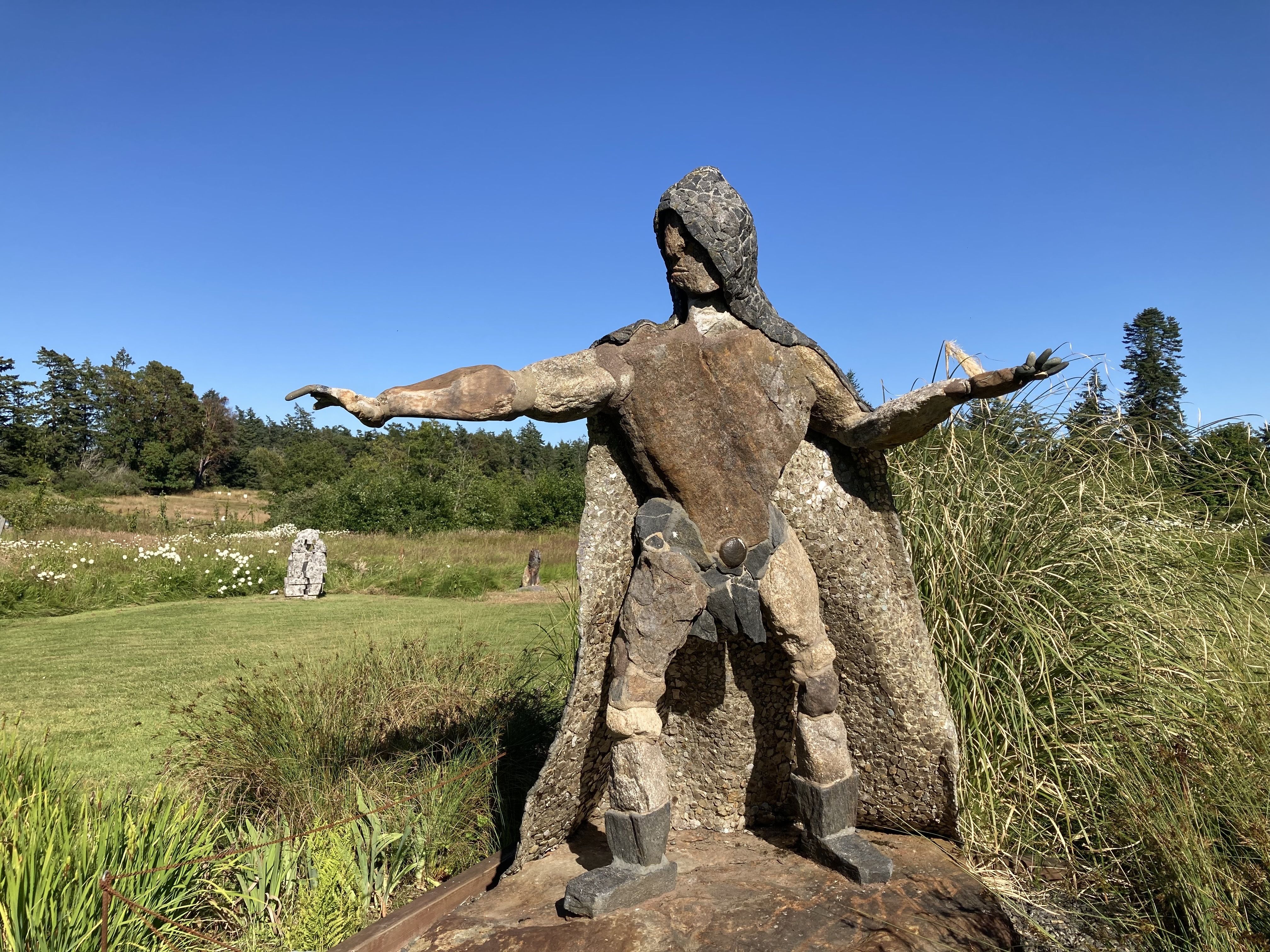 A hooded stone-and-metal human sculpture stands with outstretched arms in a grassy sculpture park beneath a clear blue sky.