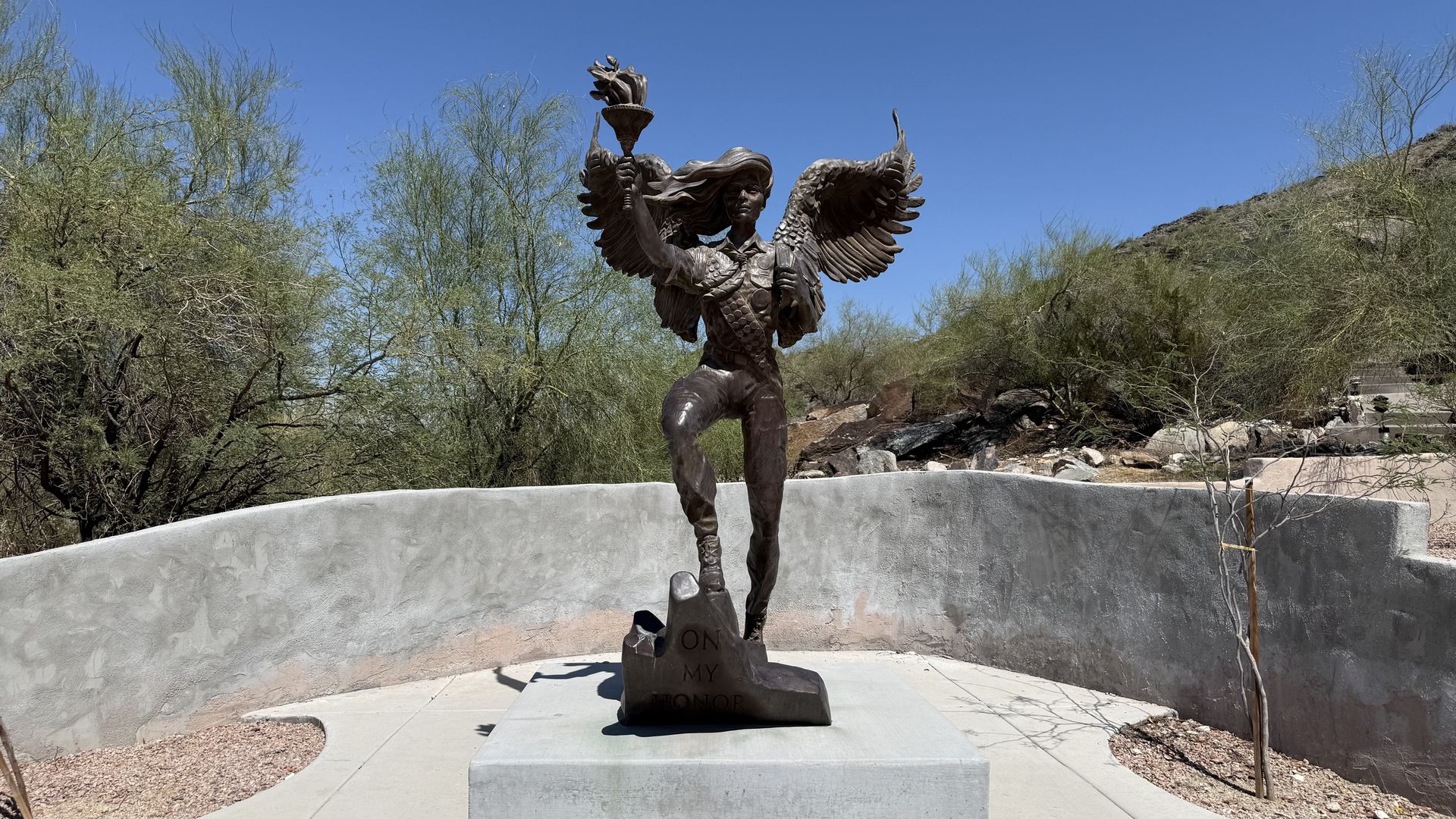 Bronze statue of a winged female warrior holding a lit torch aloft, standing on a pedestal with "ON MY HONOR" inscribed. Desert shrubs and a blue sky surround a curved concrete monument base.