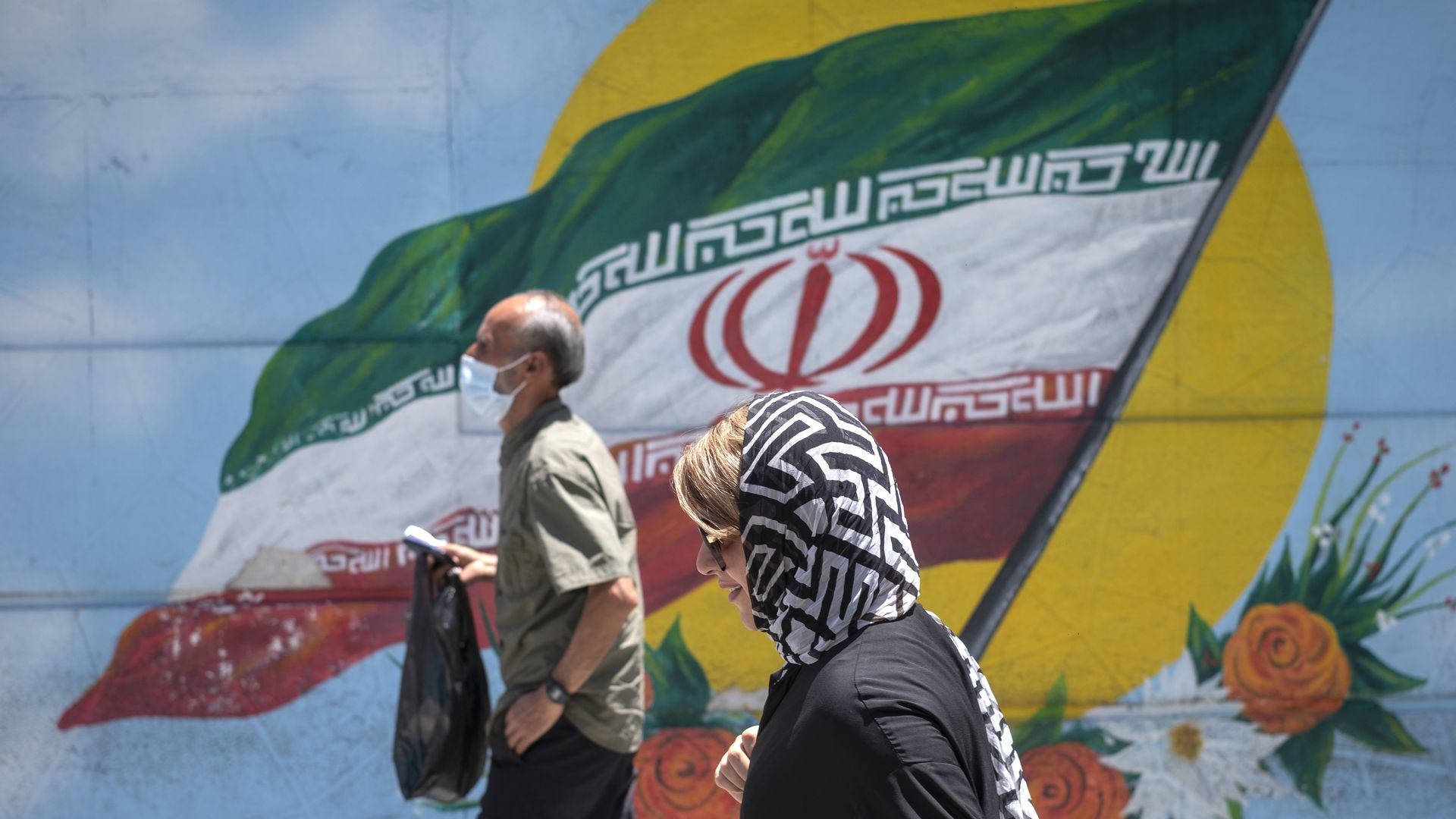 A man and a woman walk in front of an Iranian flag