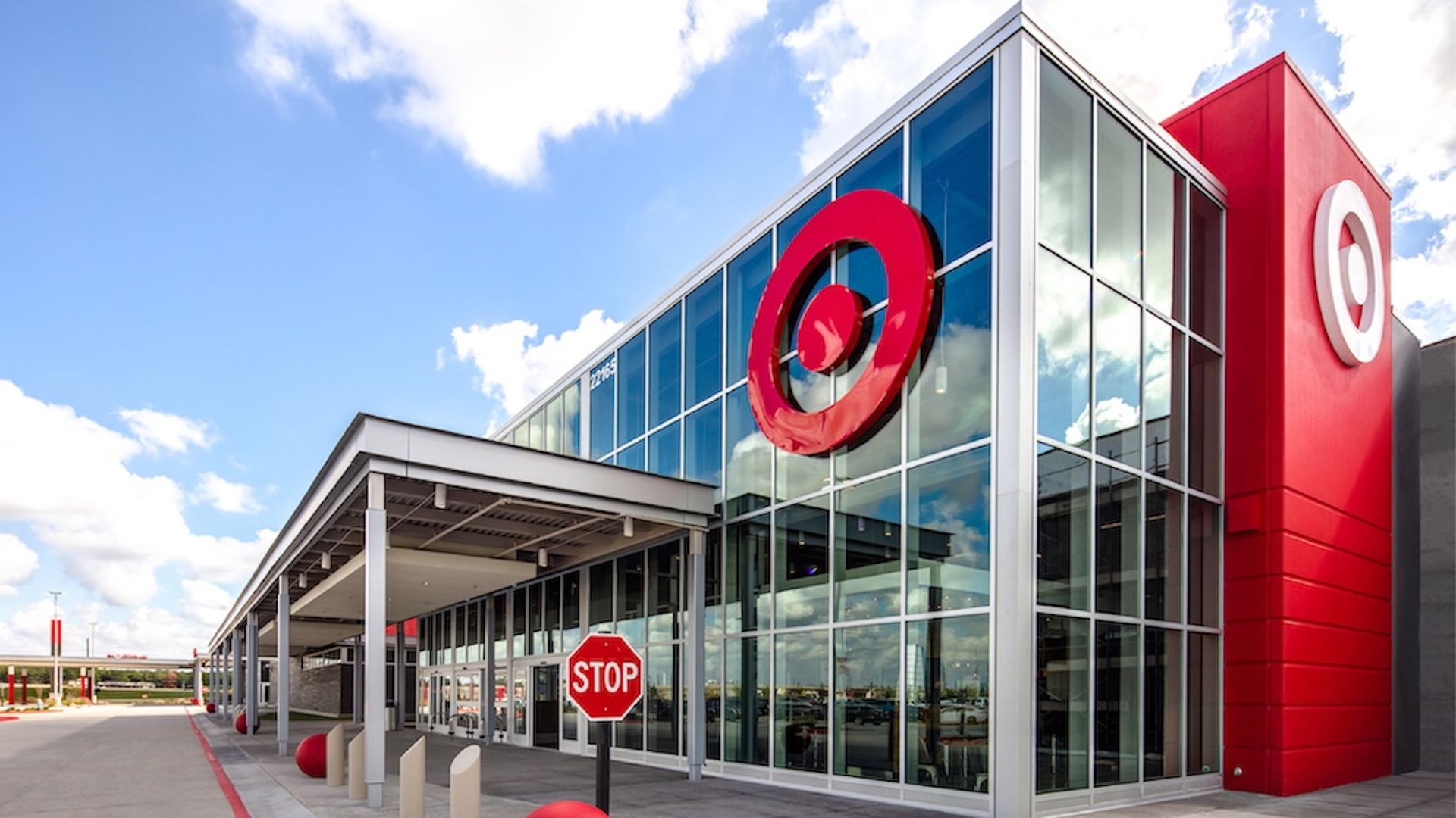 Modern glass-front Target store with large red Target logos, red spherical bollards, a stop sign, and a covered entrance under a partly cloudy blue sky.