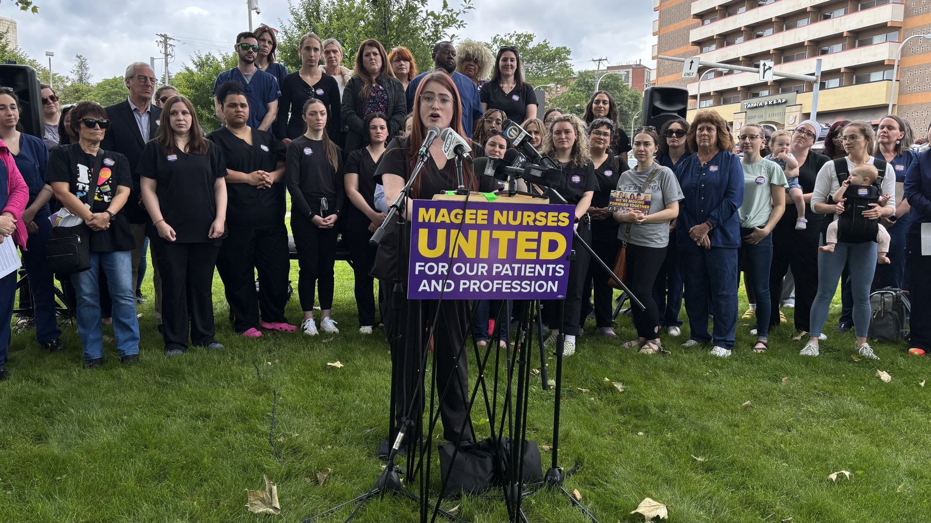 Group of nurses and supporters gathered outdoors on grass, woman in black scrubs speaking at podium with purple sign reading "Magee Nurses United for Our Patients and Profession."