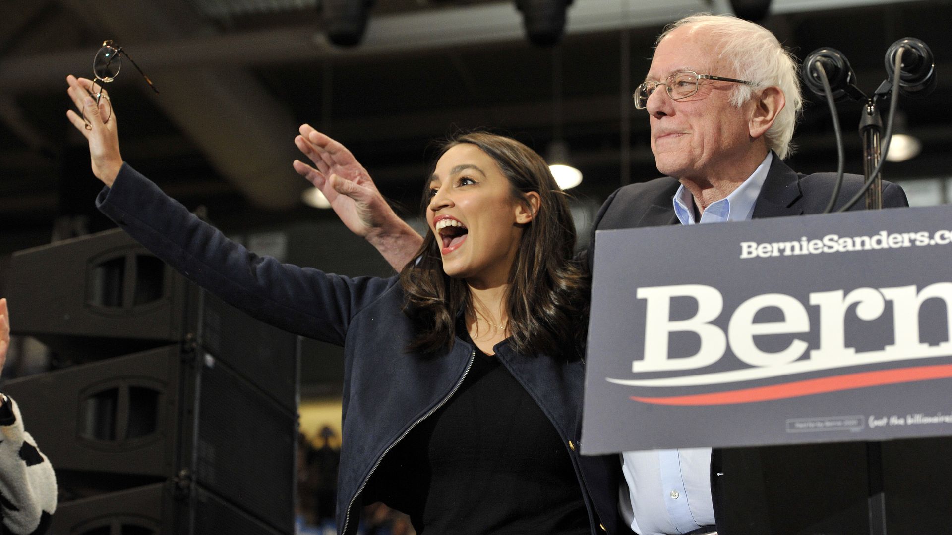 In this image, Bernie Sanders and AOC stand in front of a crowd with a Bernie 2020 sign in front of them attached to a podium.