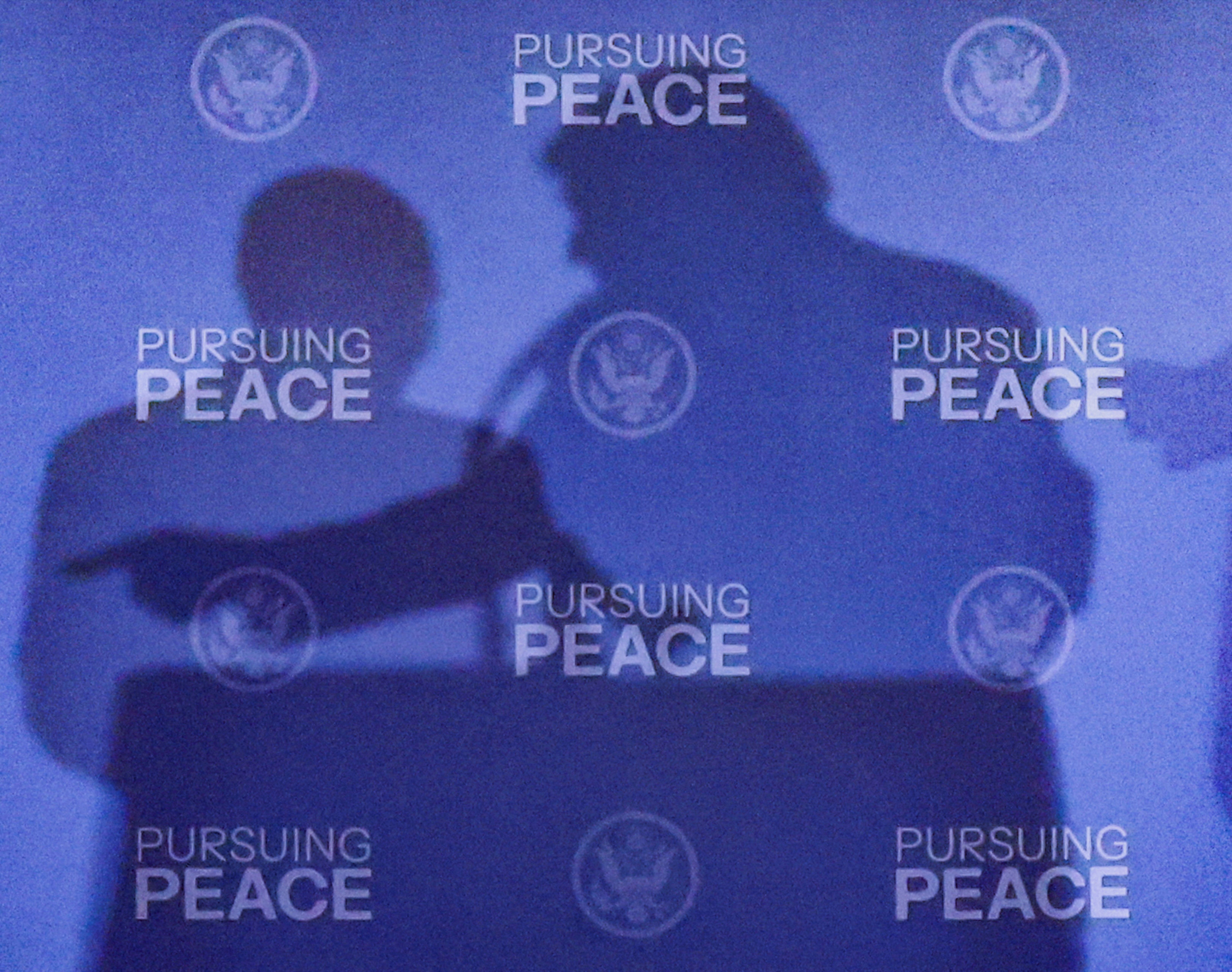 Silhouettes of two people against a blue backdrop with repeated text "Pursuing Peace" and U.S. government seals.