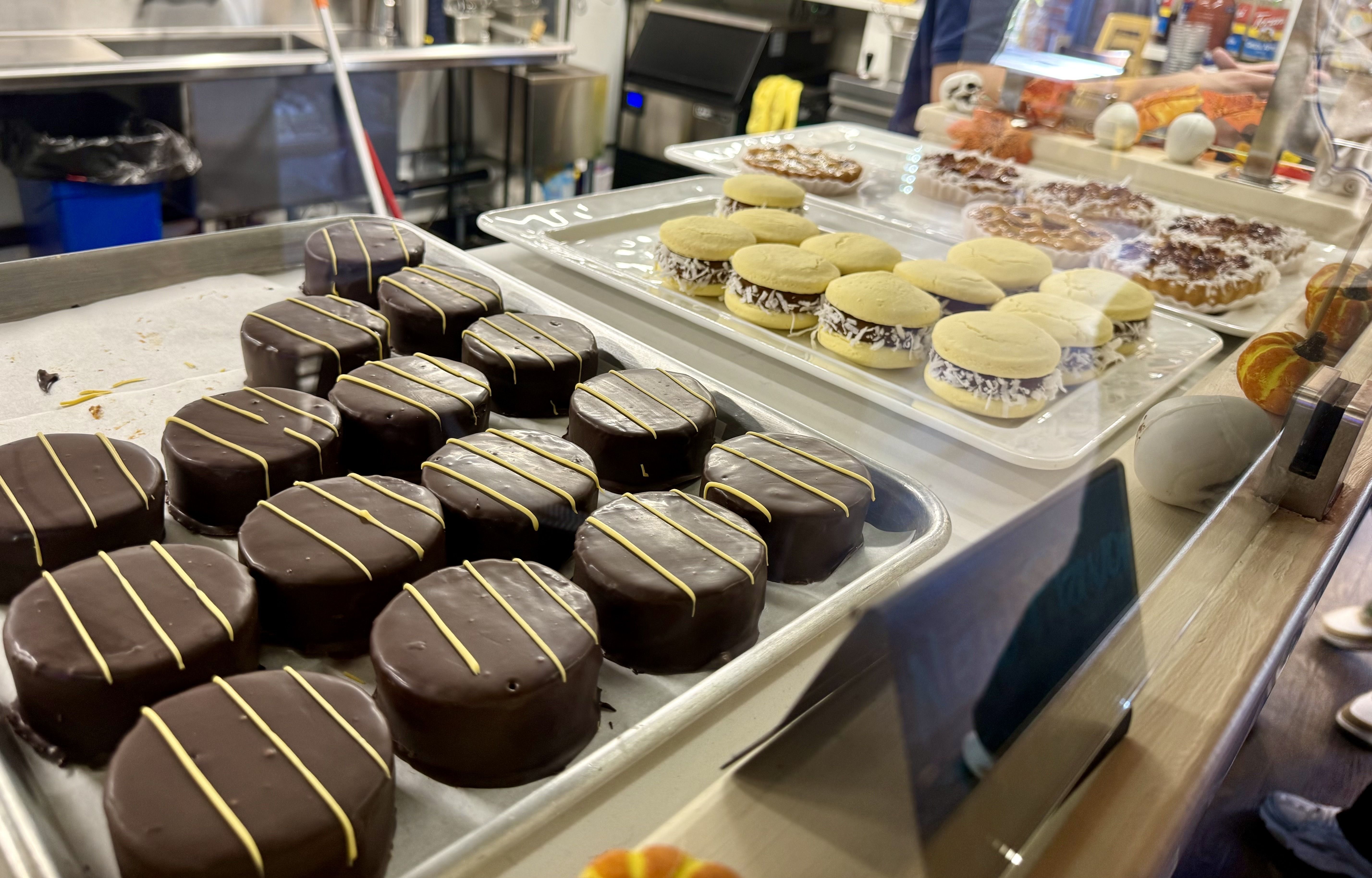 Bakery display case with dark chocolate cakes drizzled in yellow in front, beside pale yellow macarons with cream filling on trays; pastries visible in the background.