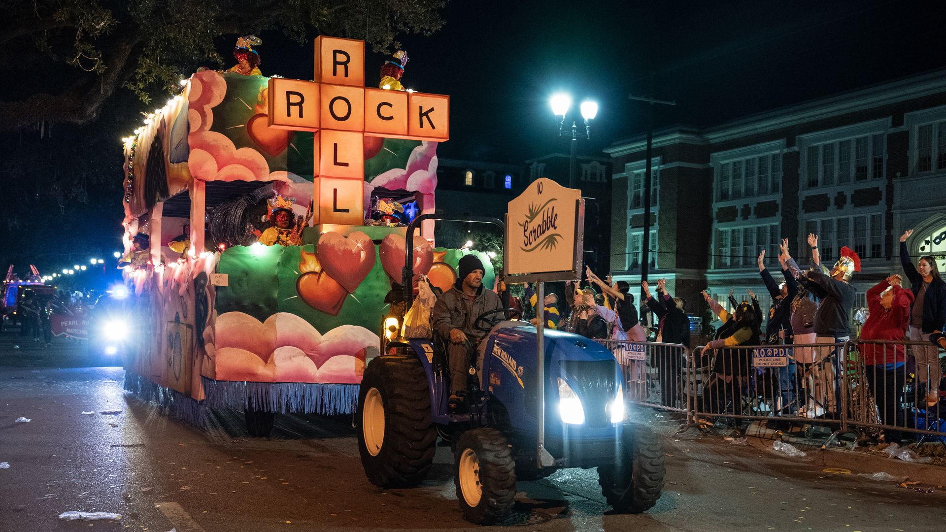 A tractor pulls a parade float, which is decorated with large Scrabble pieces spelling the words "Rock" and "Roll." Behind barricades are light crowds.