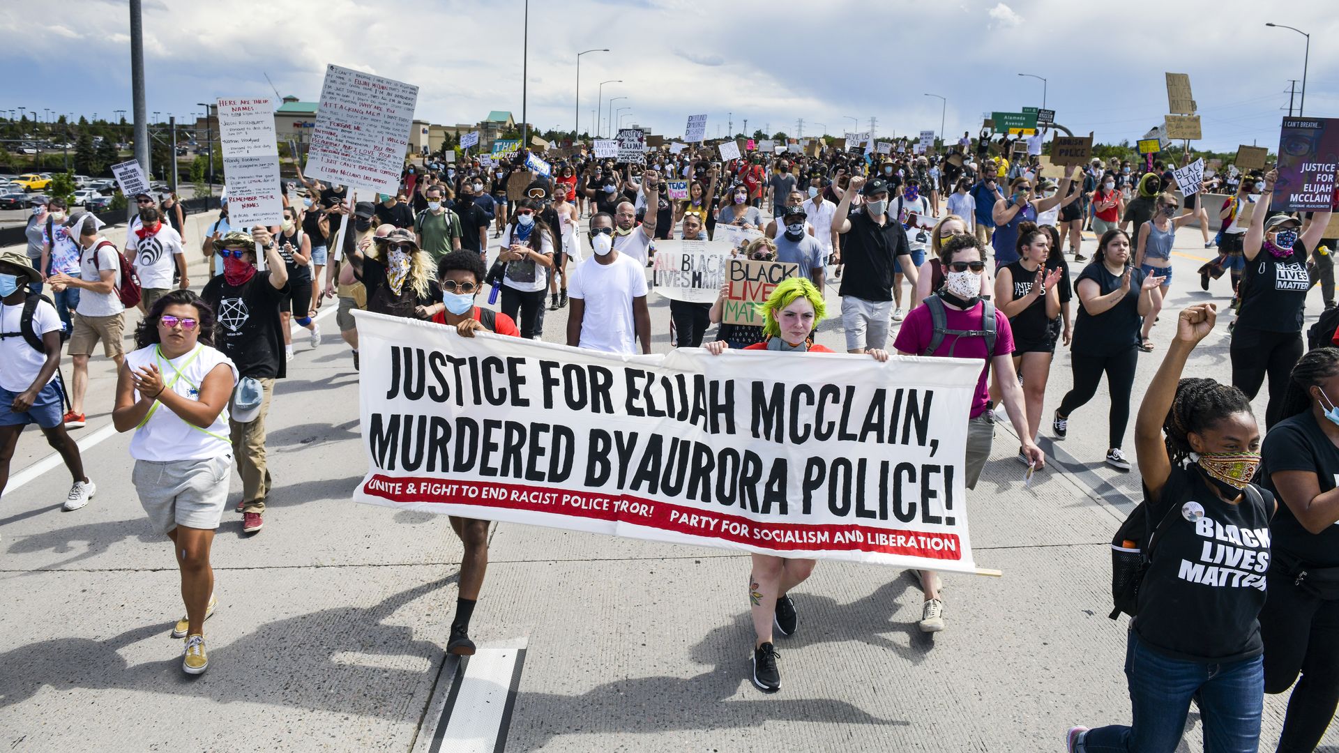 Photo of a crowd of protesters, including two holding a banner that says "Justice for Elijah McClain, murdered by Aurora police"
