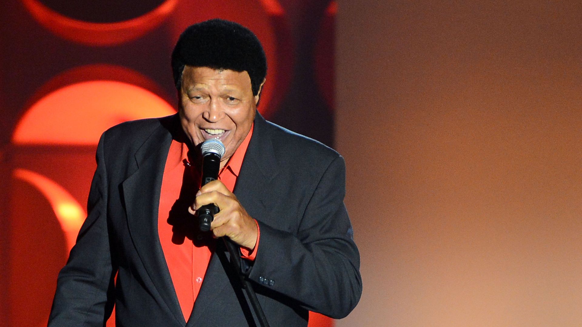 Singer Chubby Checker in a black suit and red shirt performing on stage with a microphone, red circular lights in background.