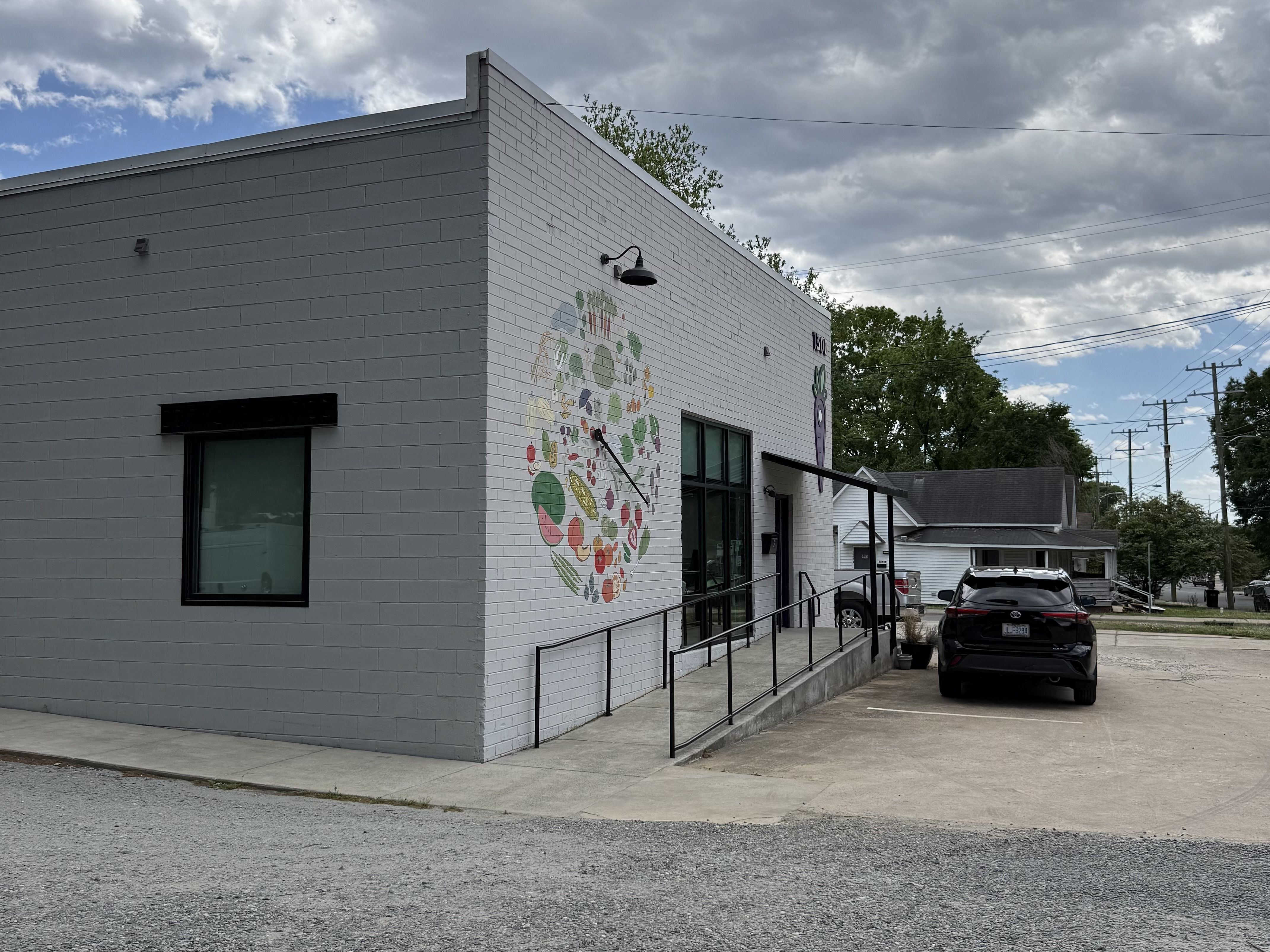Light gray brick building with a large circular, colorful mural on the side, a black outdoor lamp, and a ramp with railings. A dark SUV is parked nearby under a cloudy sky.