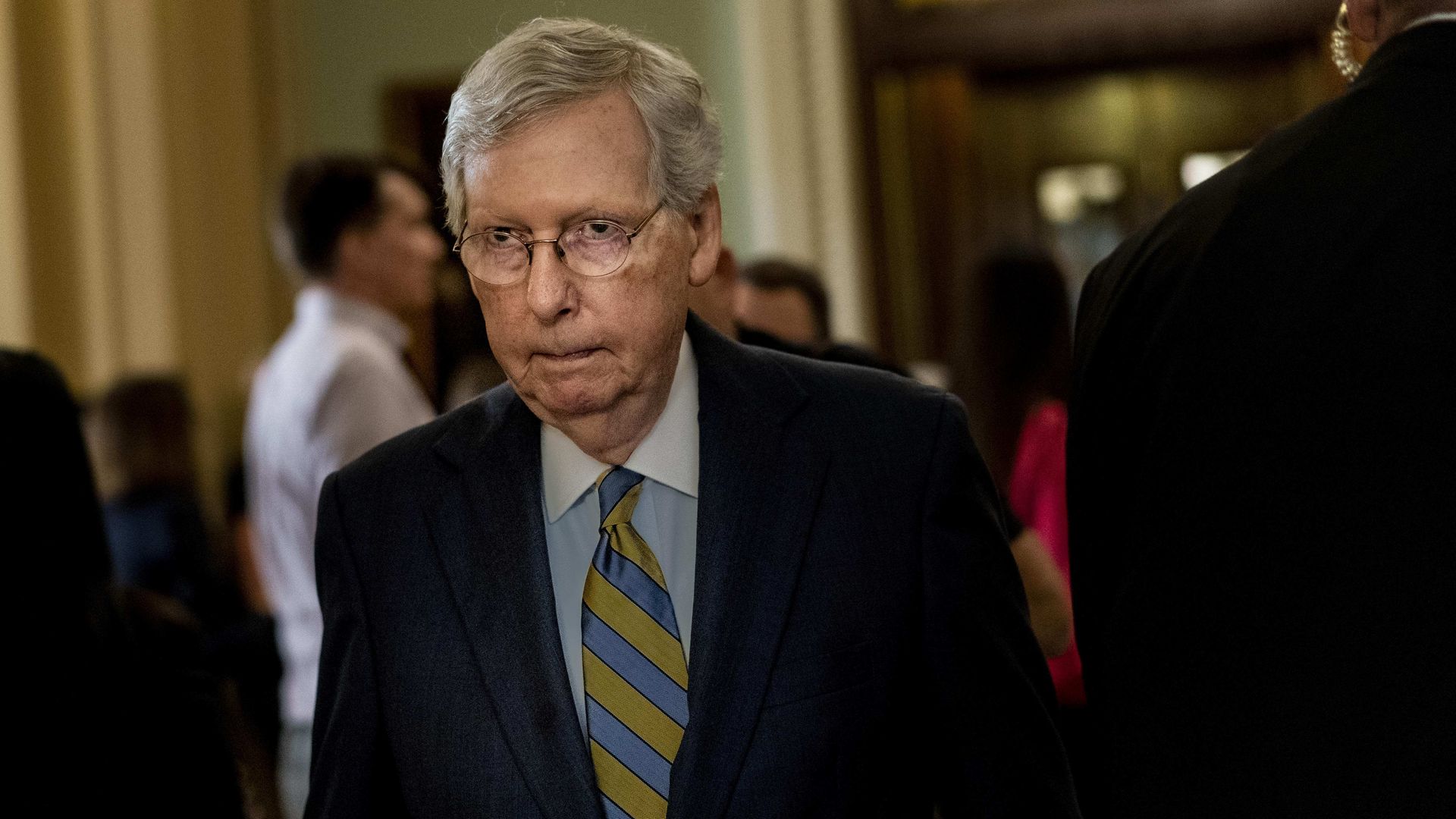 In this image, McConnell wears a stripe tie and a suit and walks through a crowded hallway.