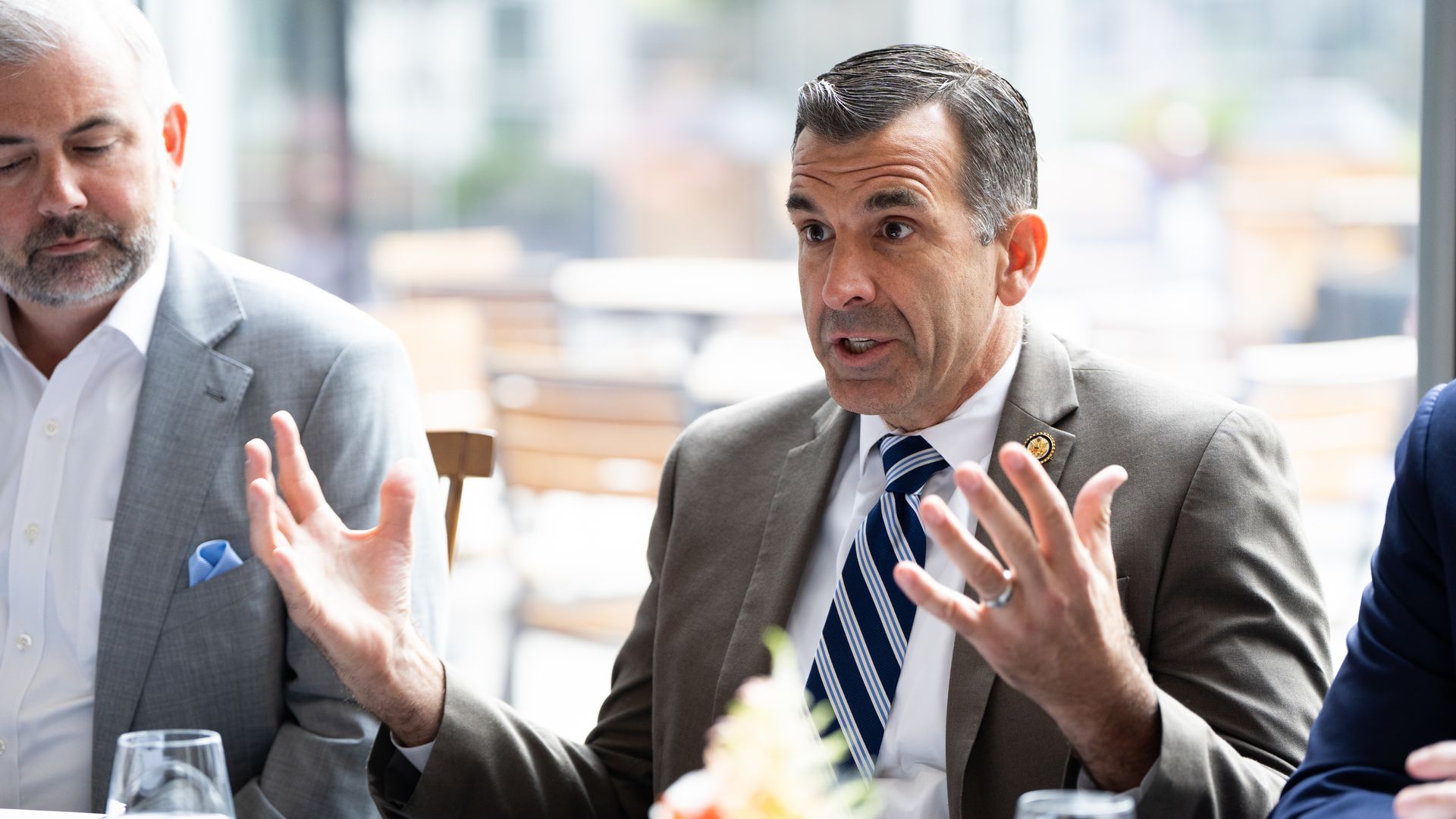 Two men in business suits debate at a glassy cafe table; the man on the right speaks with animated hand gestures, while the man on the left looks down. Water glasses and a floral centerpiece visible.