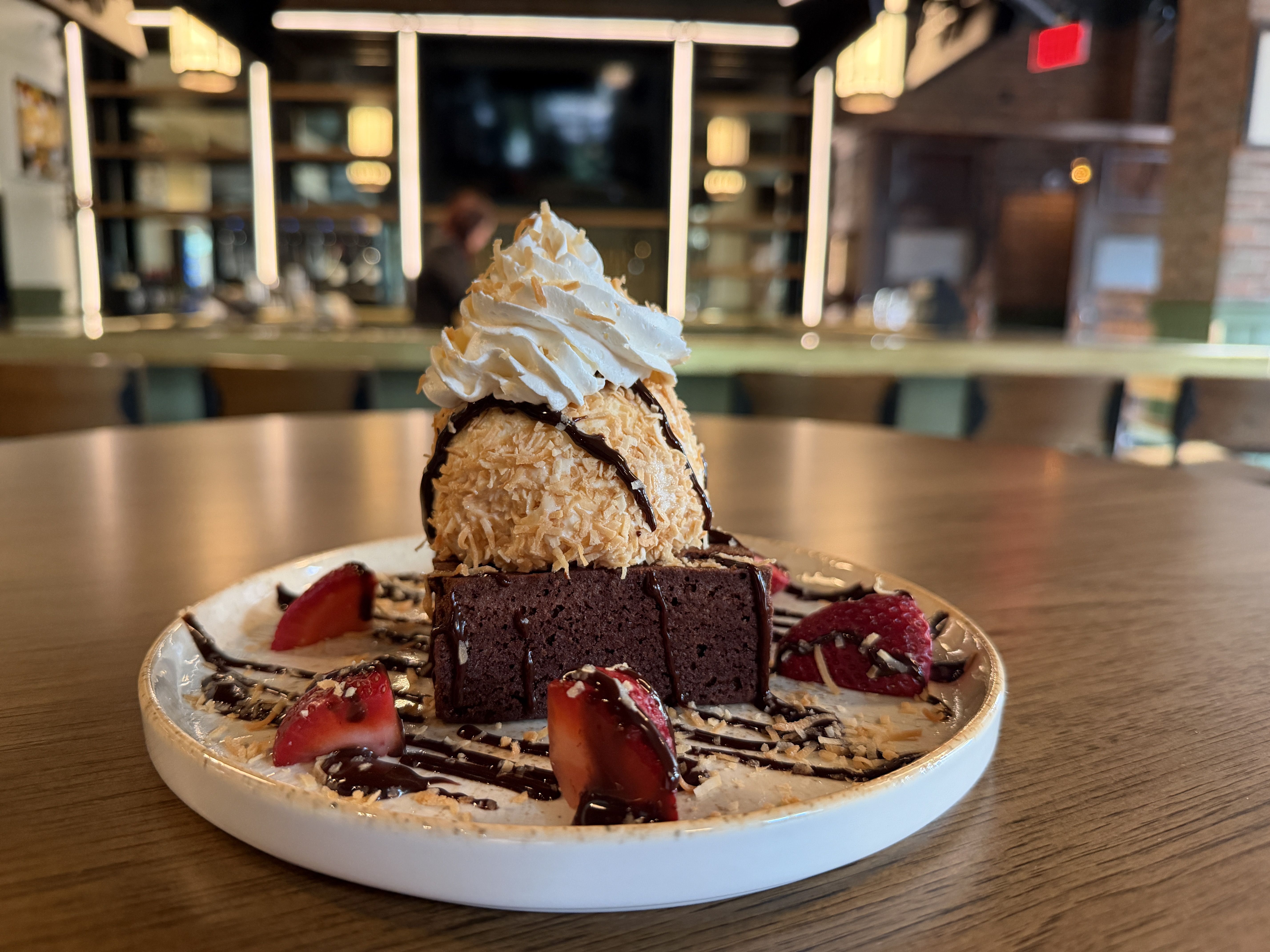 Dessert plate featuring a dark chocolate brownie base topped with a coconut-crusted ice cream scoop and a swirl of whipped cream, drizzled with chocolate. Garnished with strawberry pieces and almond slivers in a cafe.