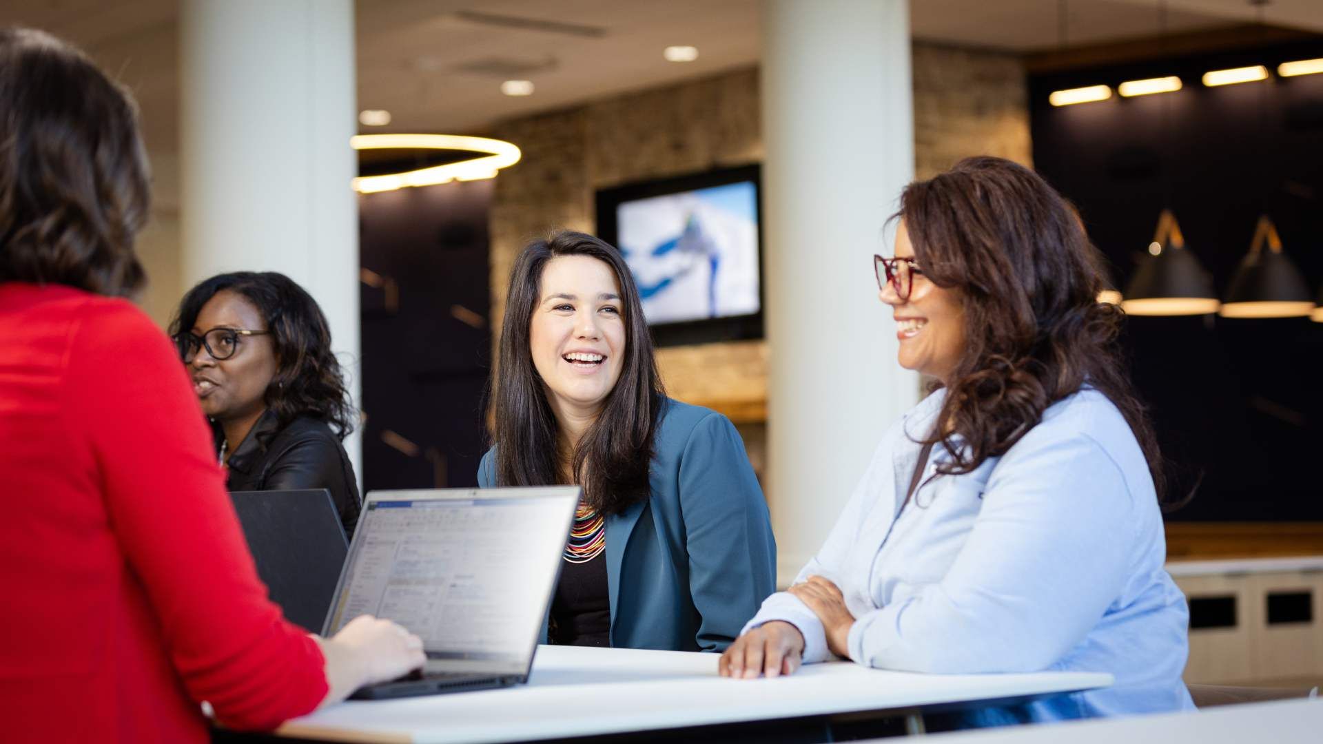 Four professional women collaborating around a table in a modern office, with laptops and warm lighting in the background.