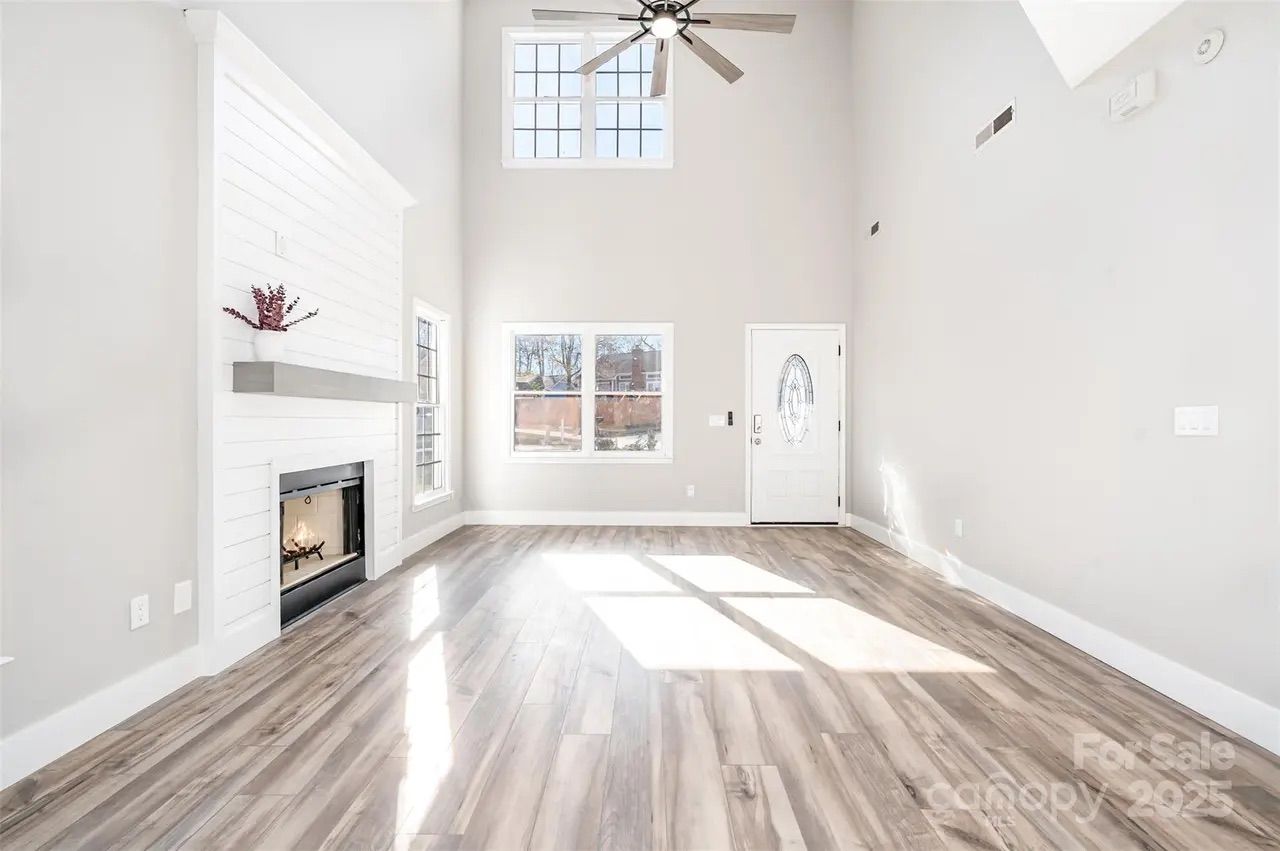 Bright, spacious living room with high ceilings, light gray walls, wood-look floor, white fireplace with a shelf and plant, large windows, ceiling fan, and white front door with oval glass.