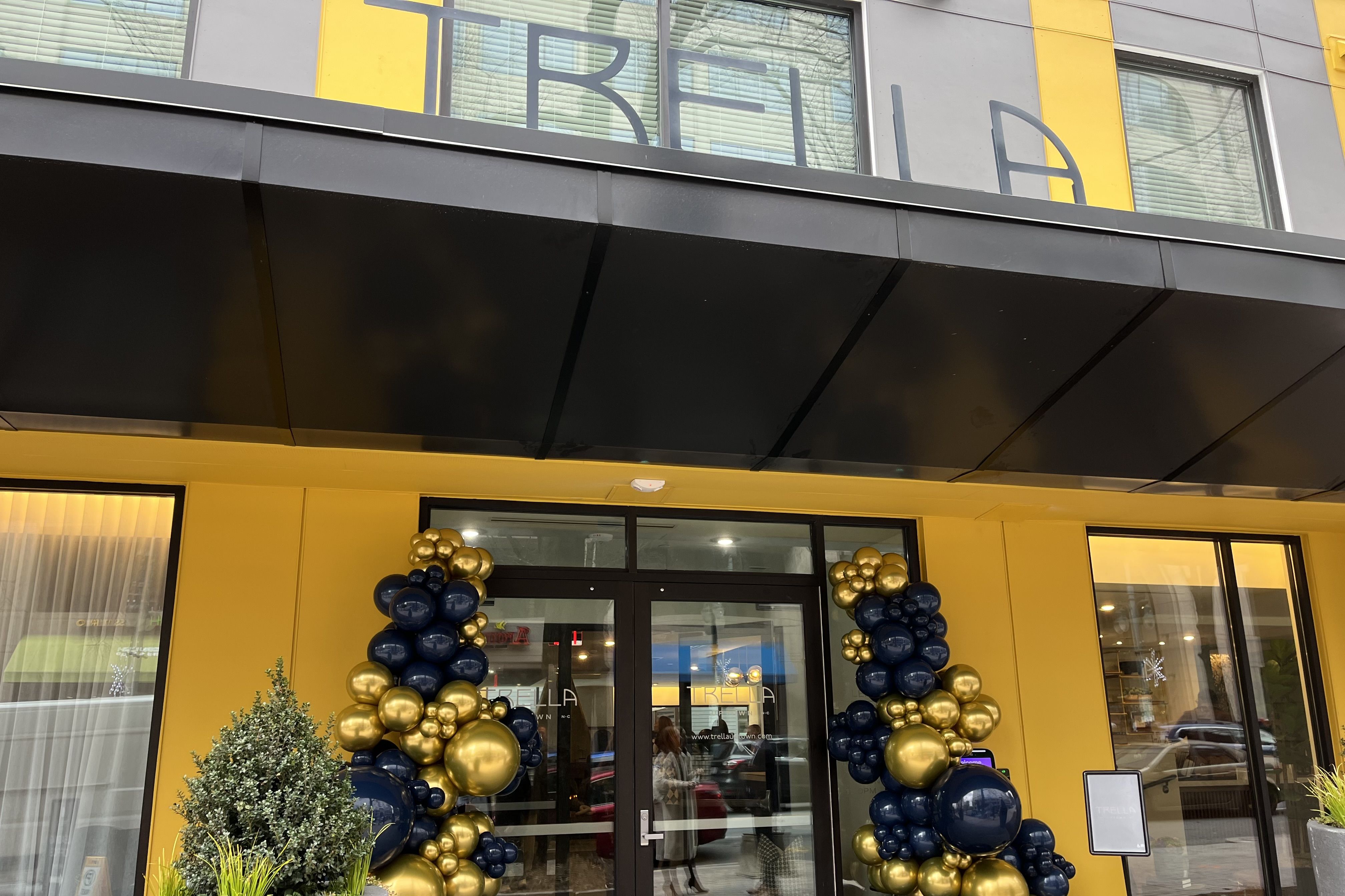Entrance to a building with bright yellow walls and black awning, decorated with black and gold balloon columns on either side of the glass door.