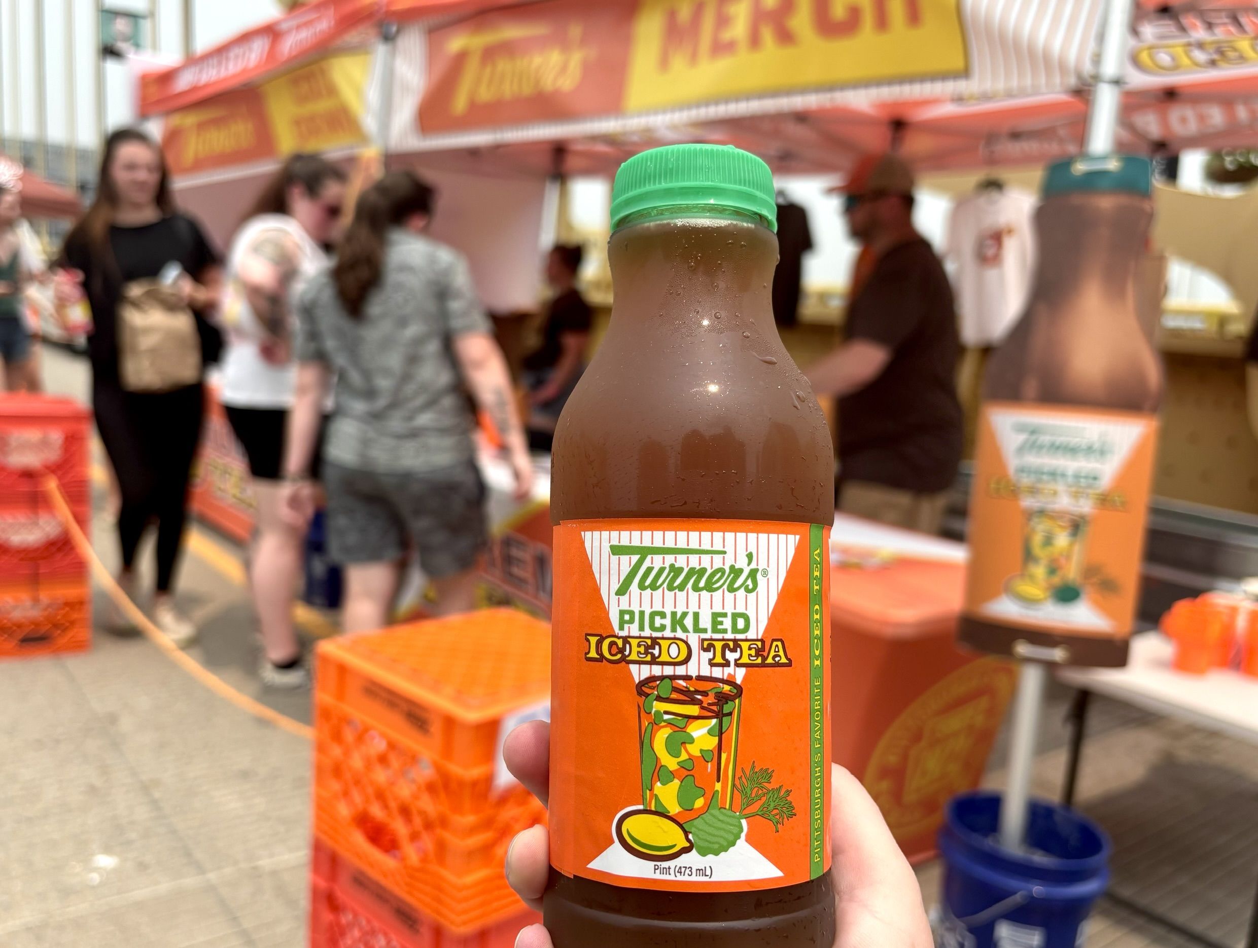 Close-up of a brown Turner's Pickled Iced Tea bottle with a bright green cap, held up in a crowded street market with orange stalls and blurred shoppers in the background.