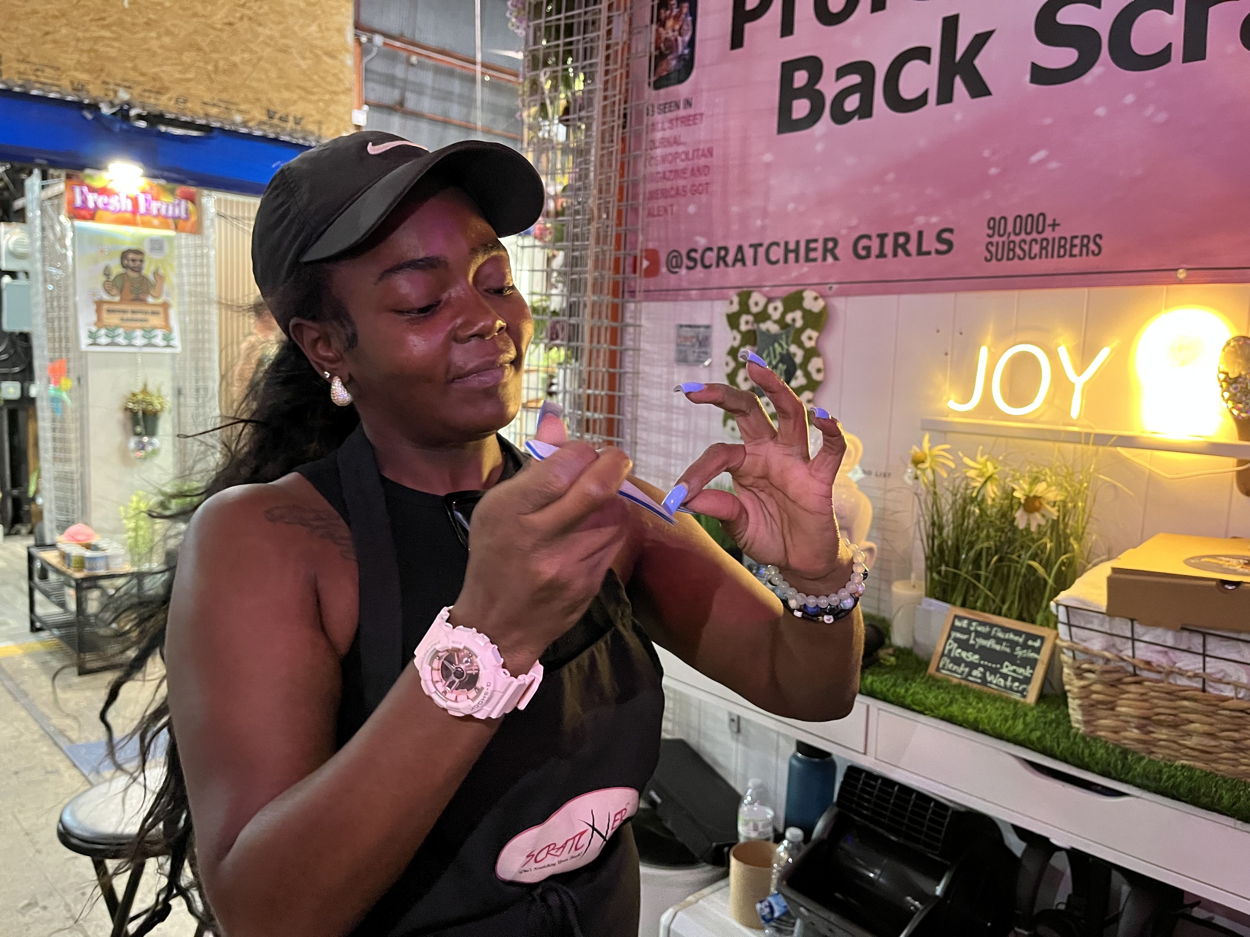Woman with a black cap and pink watch filing her nails blue at an outdoor nail booth with pink signage and a neon "JOY" sign behind her.