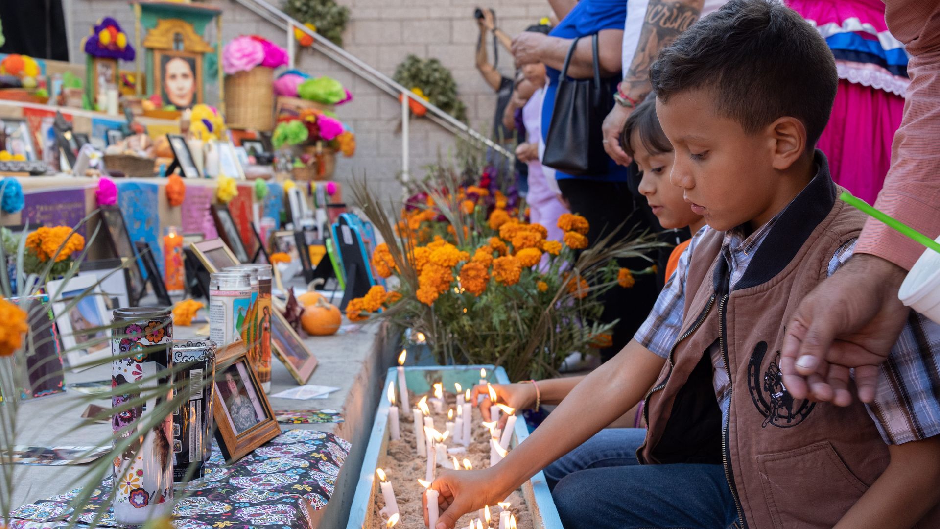 Two children lighting white candles in sand near a colorful altar with framed photos, marigold flowers, and decorations during a remembrance event.