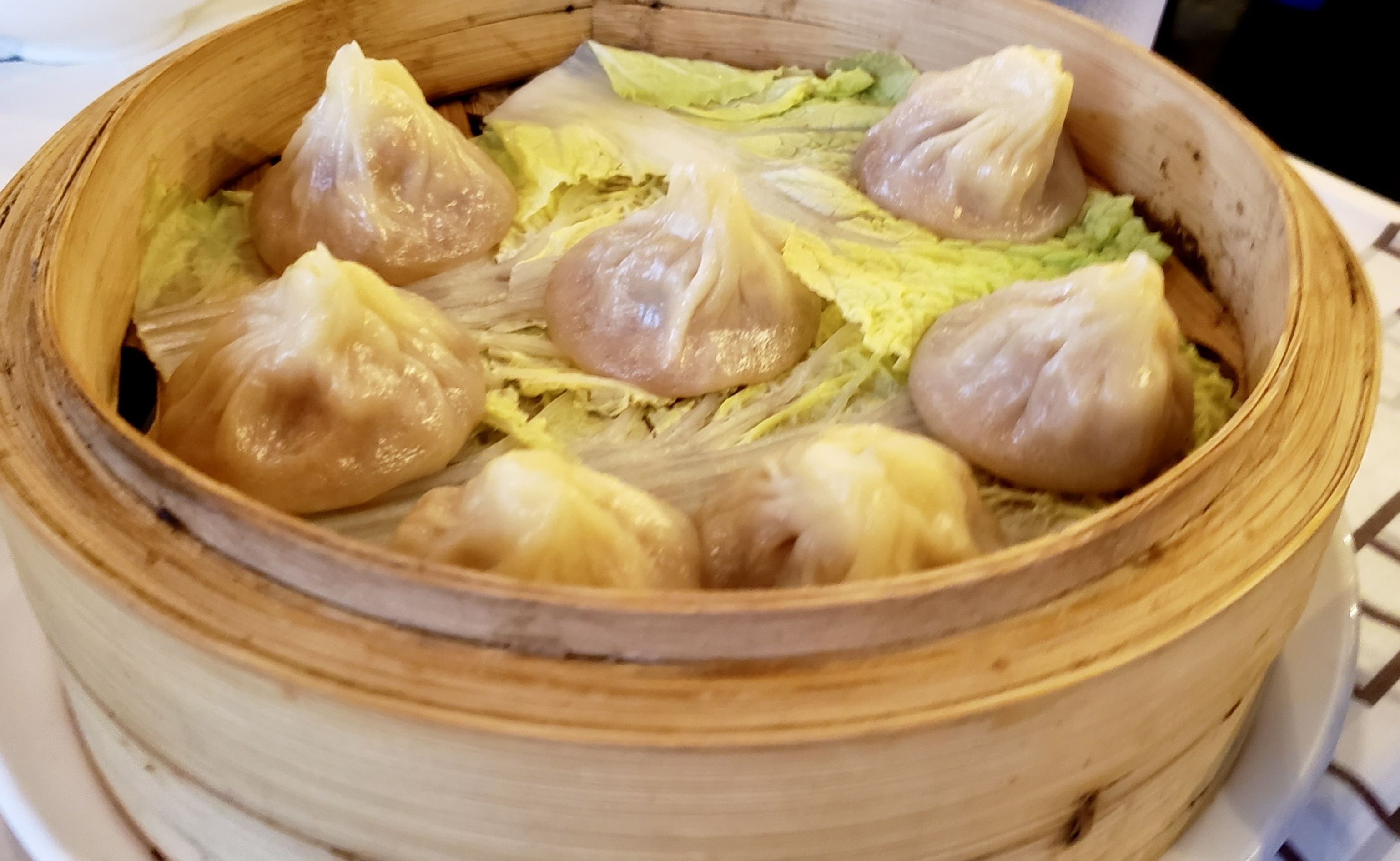 Seven steamed dumplings in a wooden bamboo steamer lined with leafy greens on a white plate.
