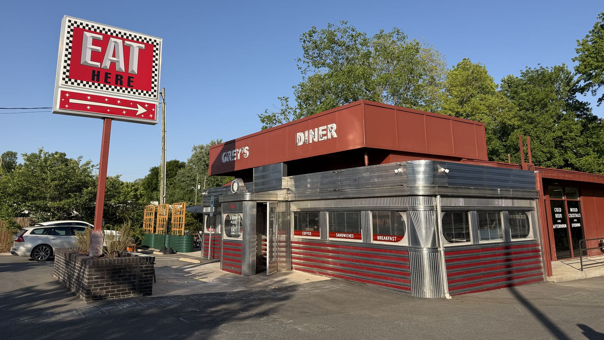 Bright blue sky over a retro diner with a red exterior and shiny metal siding. A large sign reads "EAT HERE" in bold white letters with a black-and-white checkered border, trees behind.