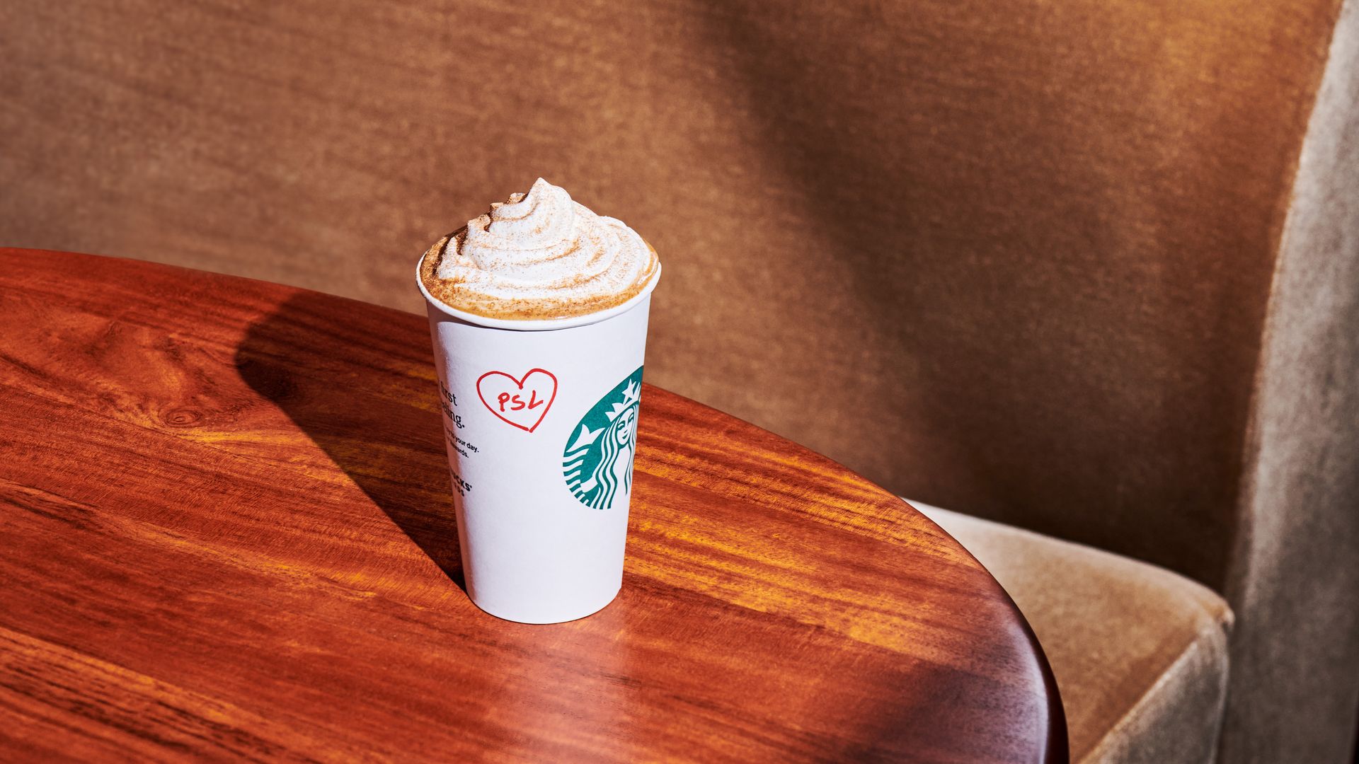 Whipped cream-topped pumpkin spice latte in a white Starbucks cup with a red heart and "PSL" written on it, sitting on a wooden table with a brown cushioned chair in the background.
